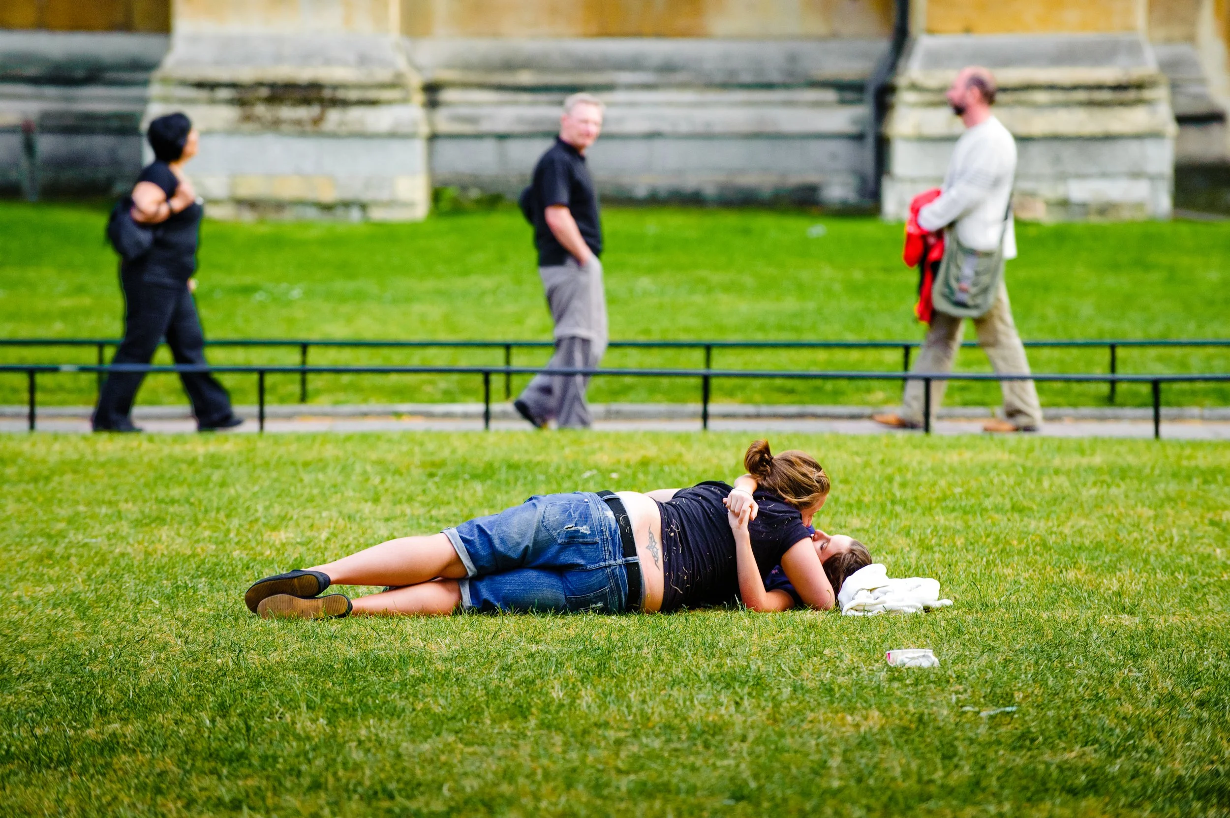 A young couple lying on the grass and kissing in an open park, with three people walking on a path behind them.