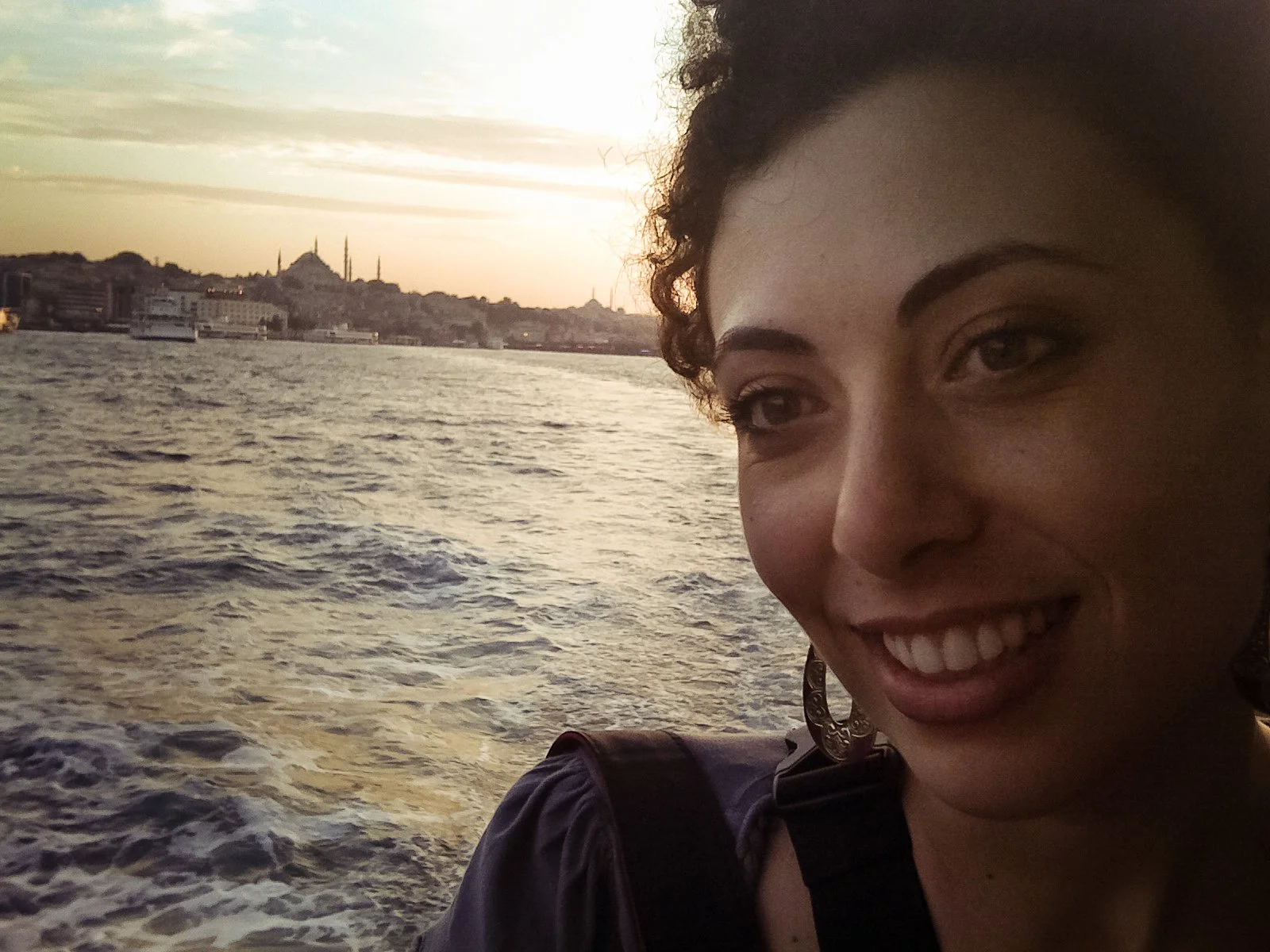 A woman with curly hair smiling on a boat, with a city skyline and a mosque in the background at sunset.