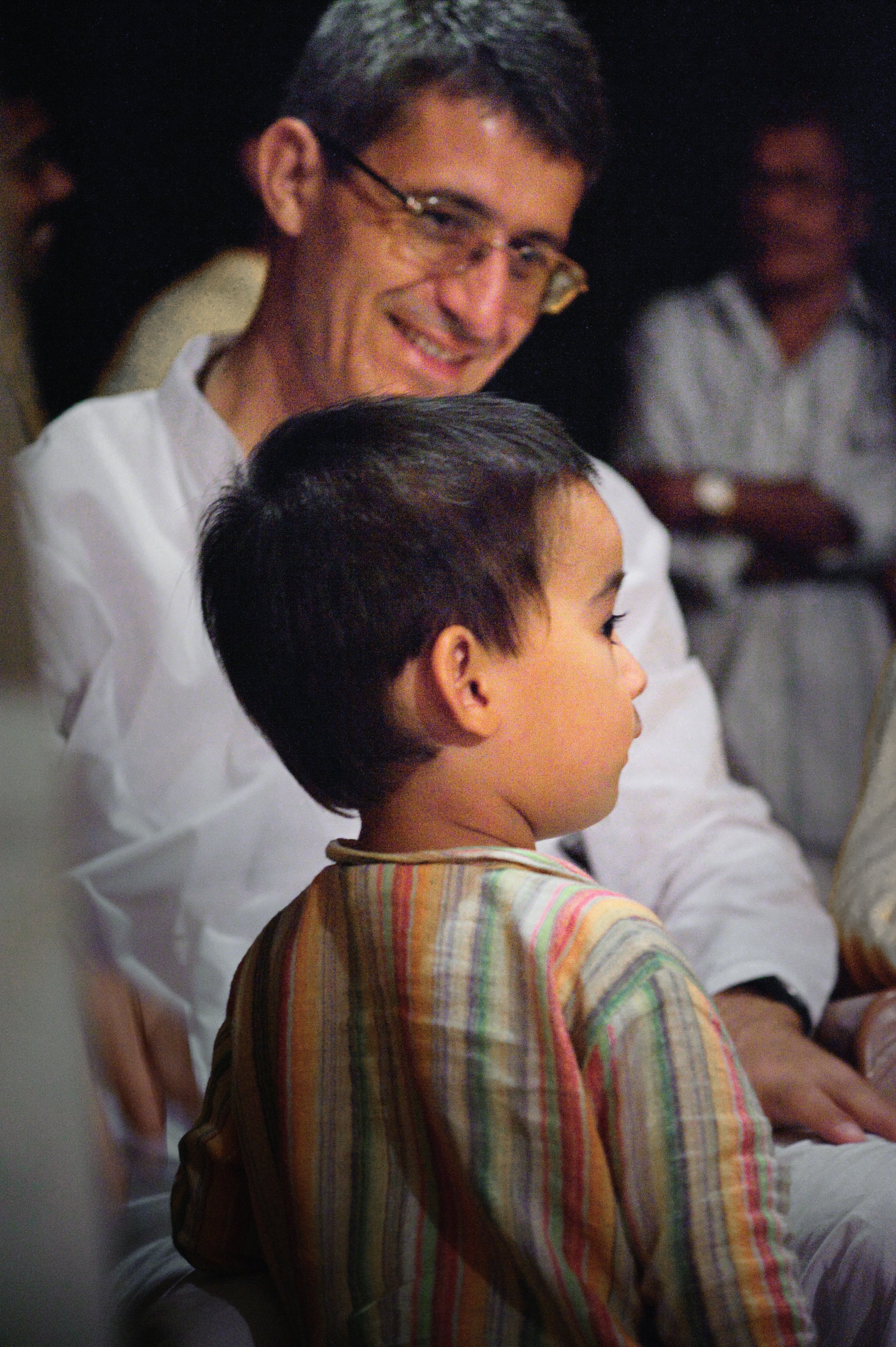 A young boy with short dark hair sitting in profile, wearing a colorful striped shirt. Behind him, an adult man with glasses and a white shirt smiling at the boy, with other people visible in the background.