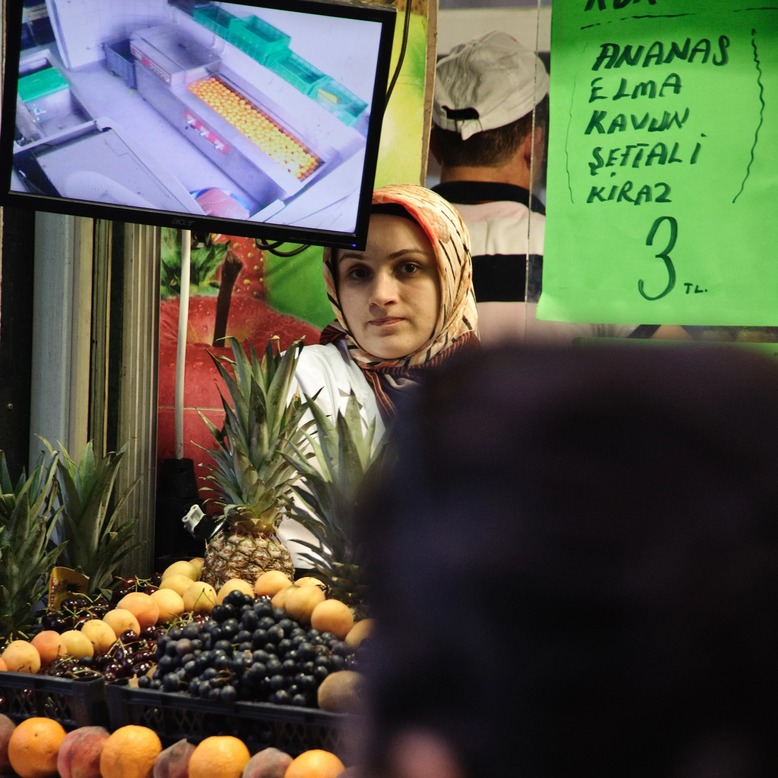 A woman wearing a headscarf looking at the camera behind a fruit stand with pineapples, oranges, peaches, and cherries. There is a green handwritten sign in Turkish listing fruit prices, and a monitor displaying a close-up of the fruit. A man wearing