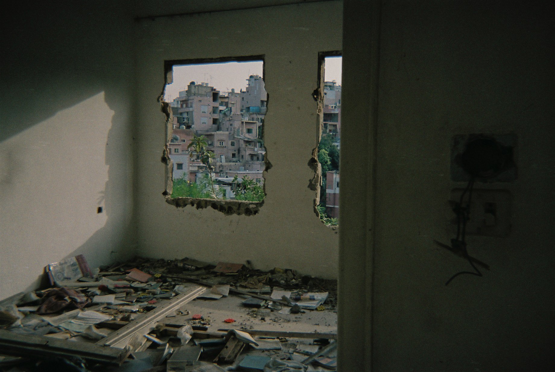 Abandoned room with damaged walls and a torn window, overlooking a densely populated hillside with numerous apartment buildings.