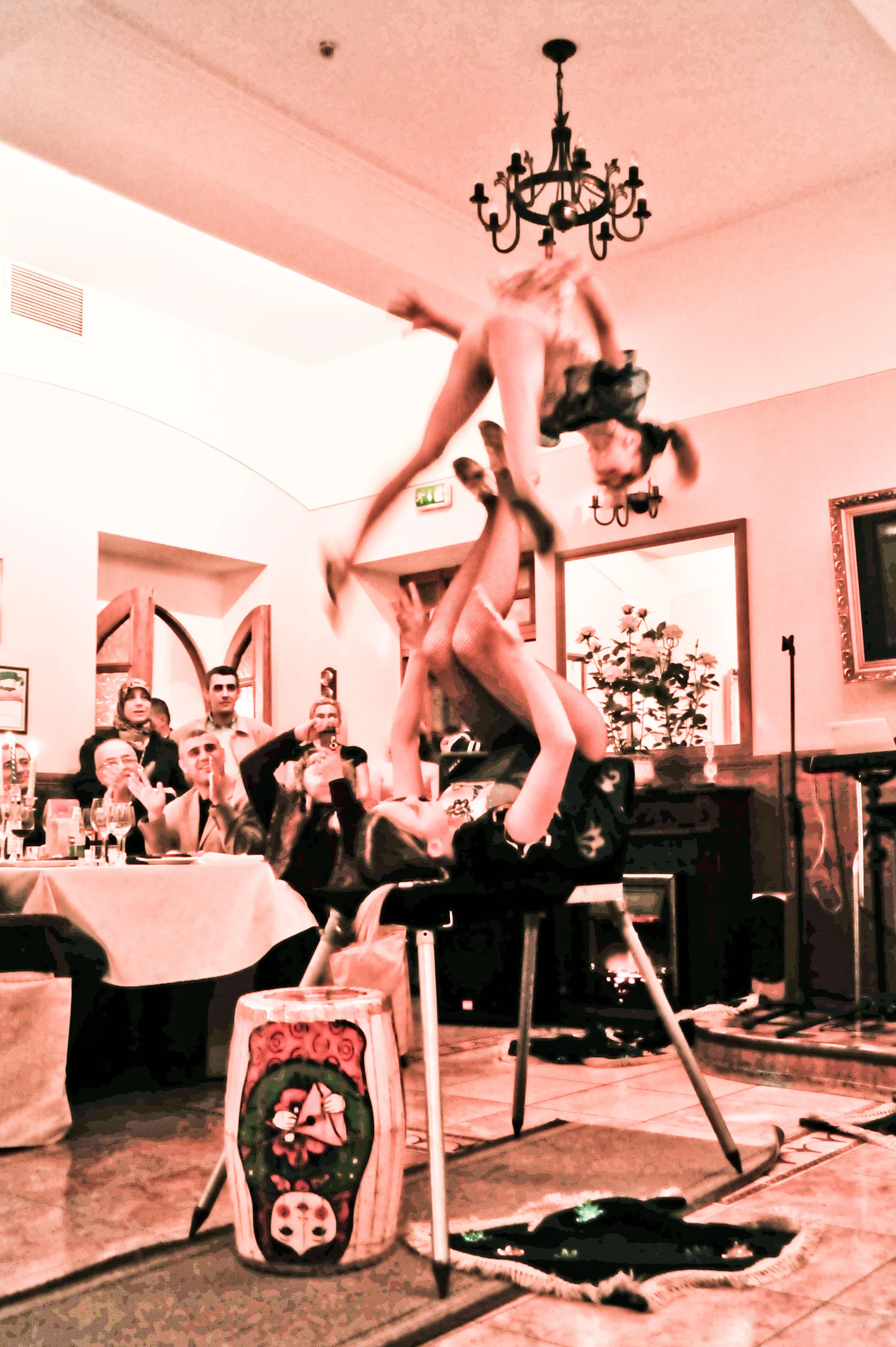A woman performing a balancing act on a chair with a man holding her up at a party or celebration, with guests watching and taking photos.