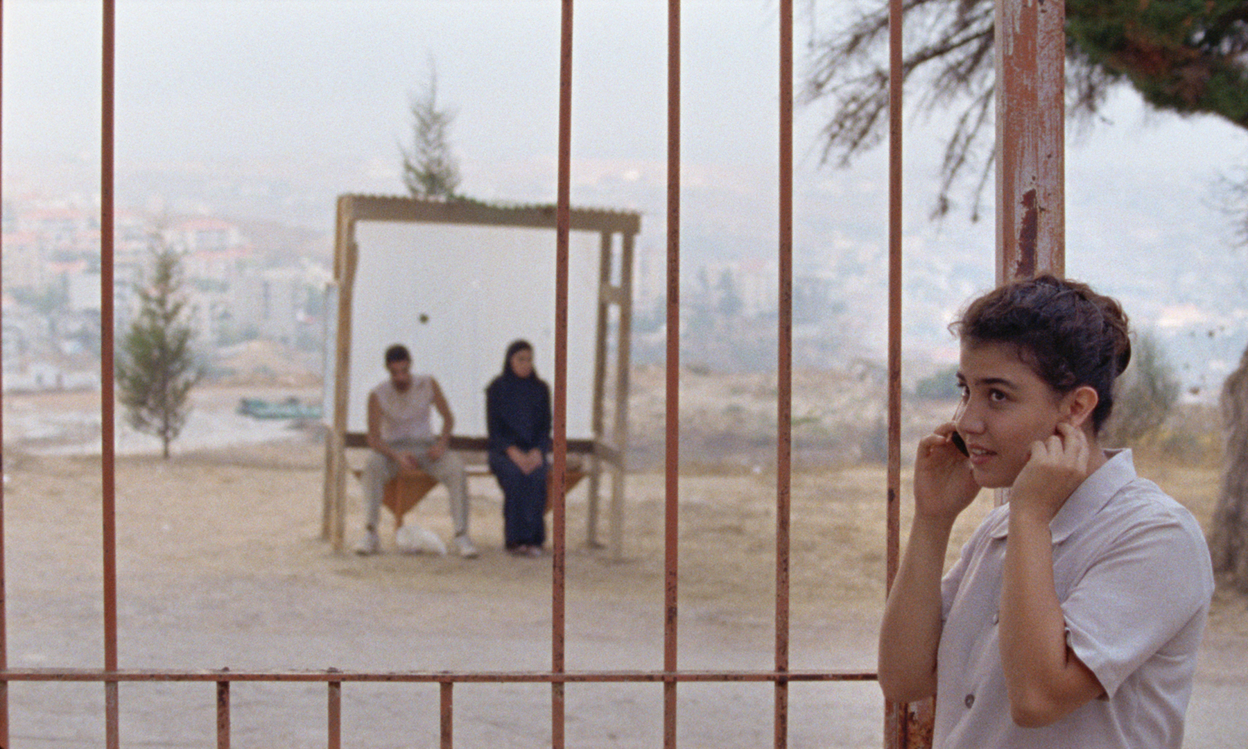 A young woman talking on a cellphone, standing behind rusted bars, with two people sitting on a bench in a blurry background.
