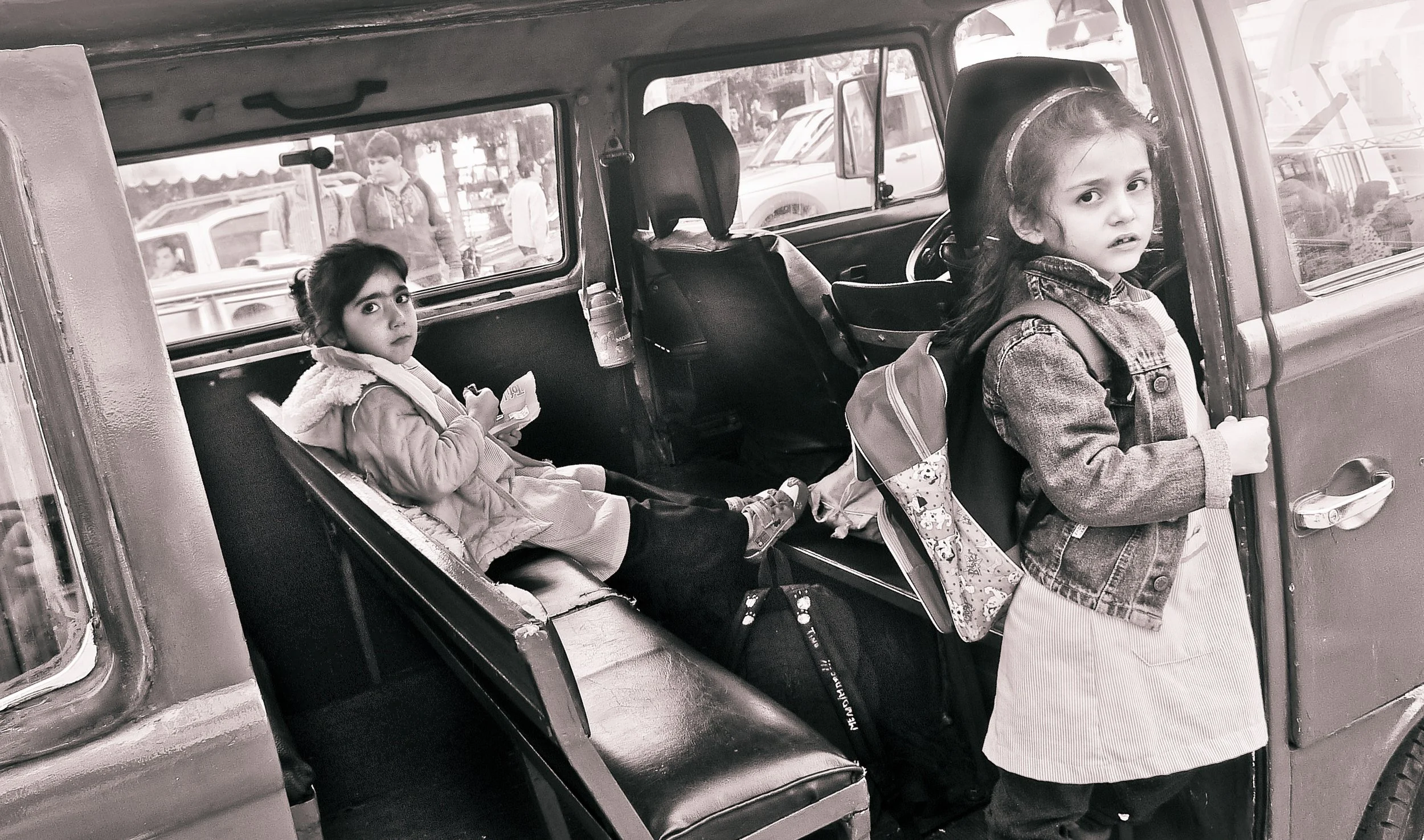 Two young girls with backpacks, one standing outside a vehicle and the other sitting inside, looking towards the camera. The vehicle appears to be an older model, and there are other cars and people visible outside.