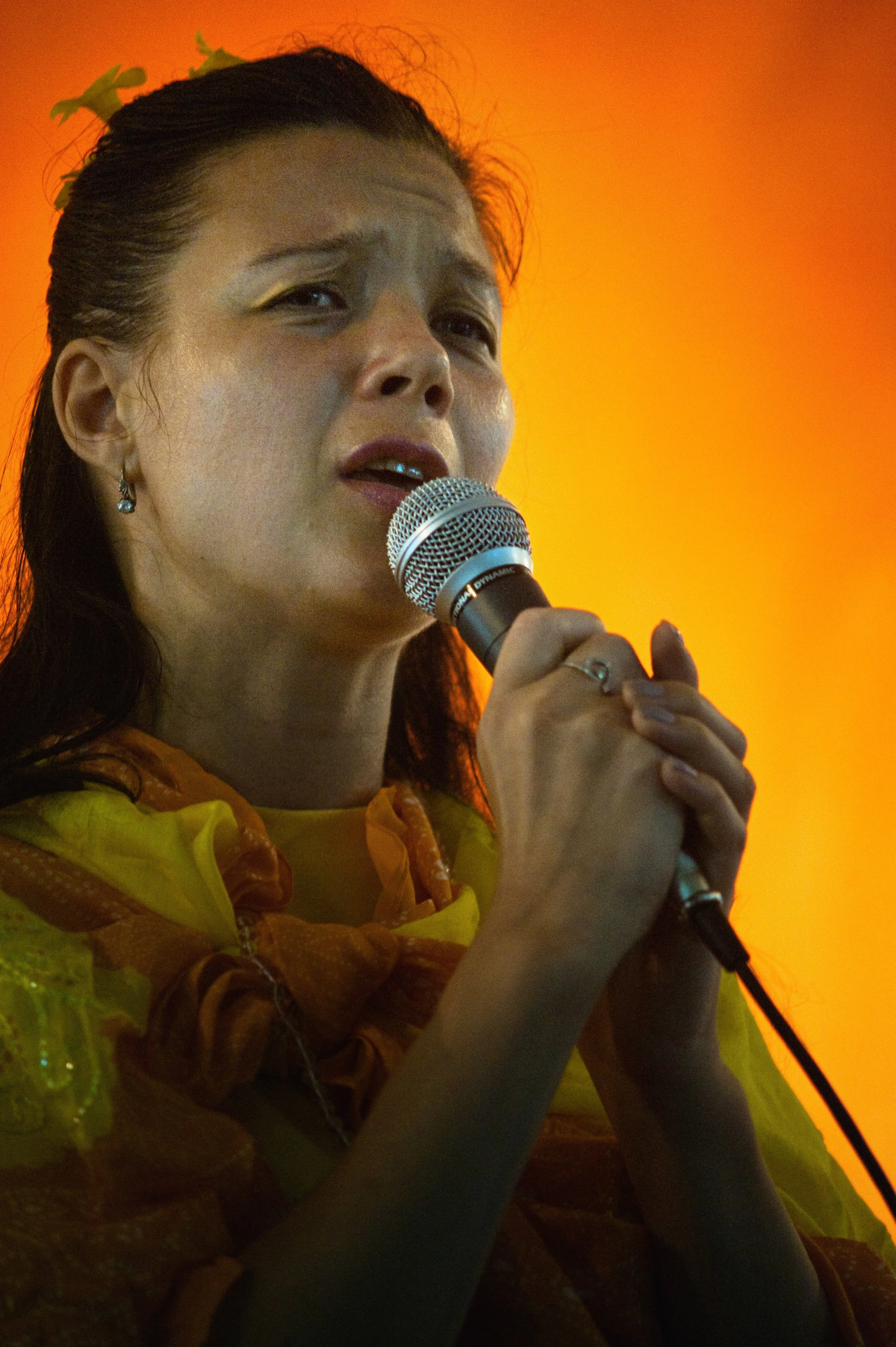 A woman singing into a microphone with a serious expression, wearing a yellow and orange outfit and earrings, with an orange background.