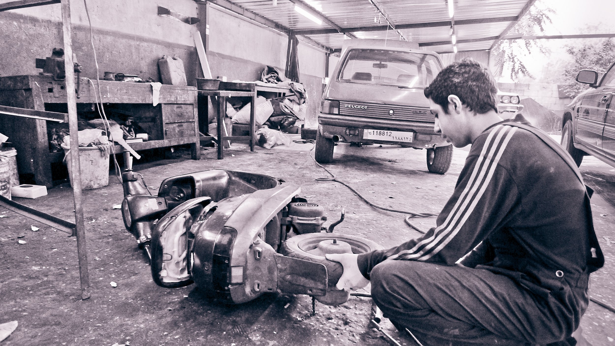 A young man in a black jacket with white stripes on the sleeves is working on a disassembled car engine in a cluttered garage, with a Peugeot van and car in the background.