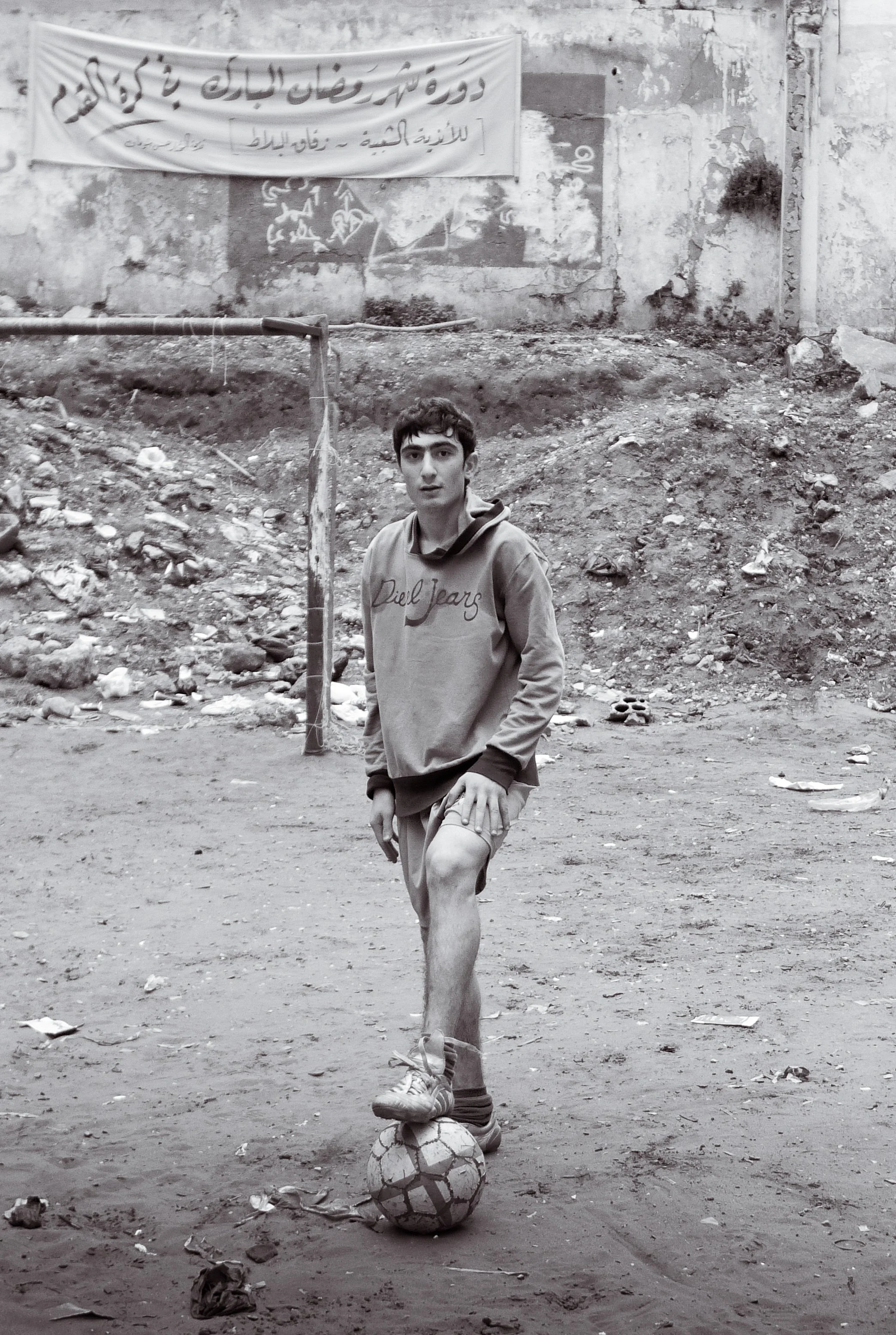A young man standing outdoors on dirt ground, balancing on one foot on a soccer ball with one leg raised, wearing a hooded sweatshirt and shorts. Behind him, there is a wall with some graffiti and a banner with Arabic writing, and a makeshift goal po