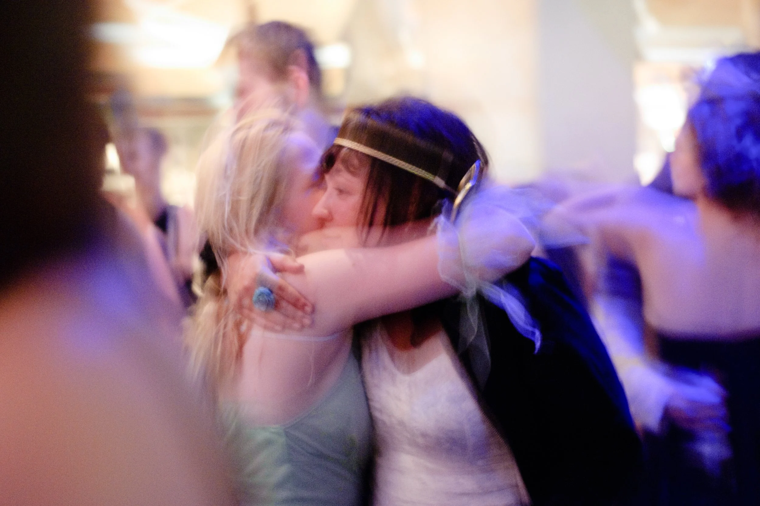 Two women embracing on a dance floor, surrounded by other people, in a lively and colorful setting.