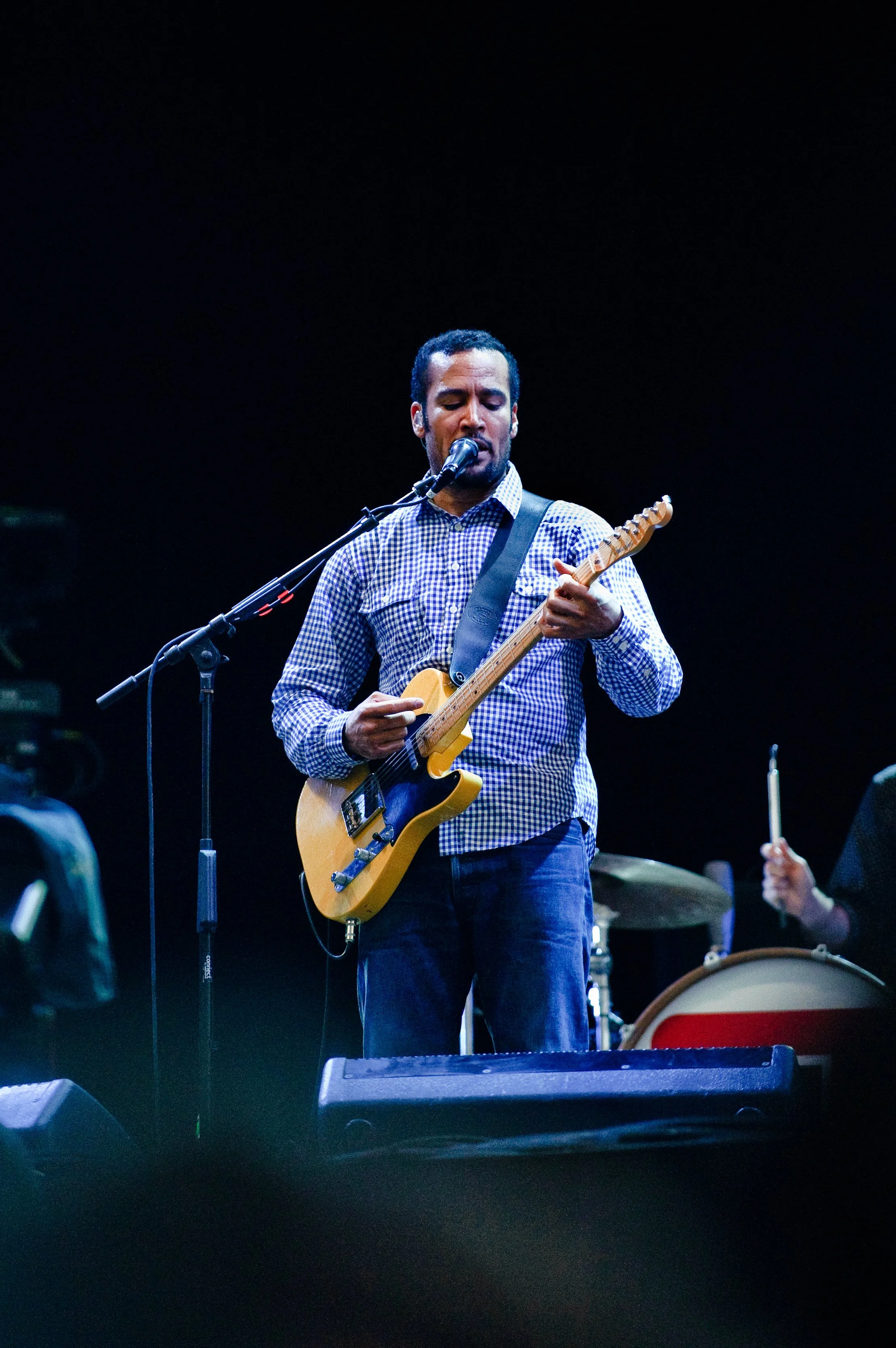 Male musician singing into a microphone while playing an electric guitar on stage with a drummer in the background.