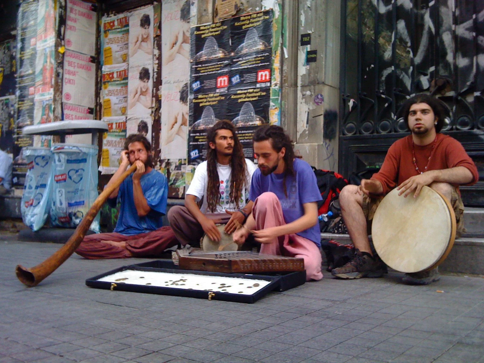 Four street musicians sitting on the sidewalk in front of a wall covered with posters. One playing a didgeridoo, another with hand drums, and the third with a tambourine. A tray of coins is in front of them.