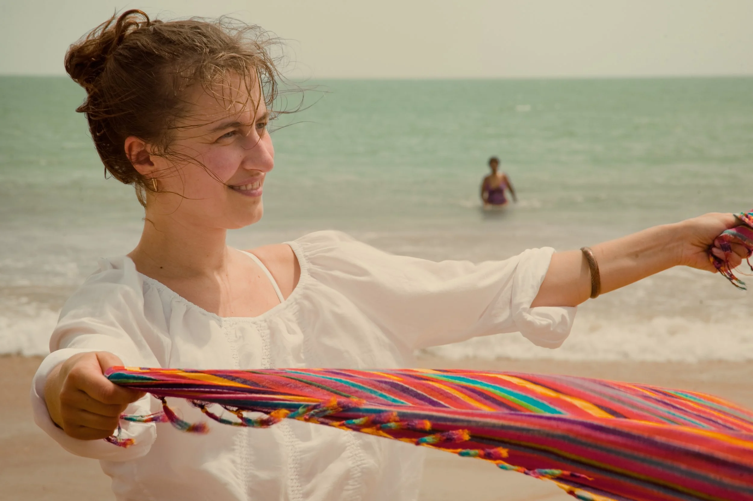 Woman on the beach holding a colorful striped fabric or blanket, smiling, with another person in the water in the background.