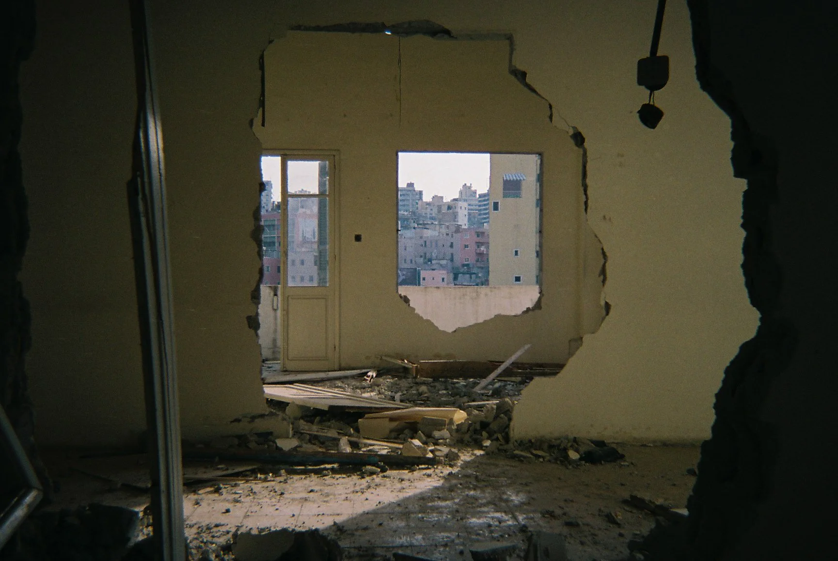 A damaged room with a large hole in the wall, exposing a cityscape through the window. Debris on the floor includes broken pieces of drywall, wood, and other materials.