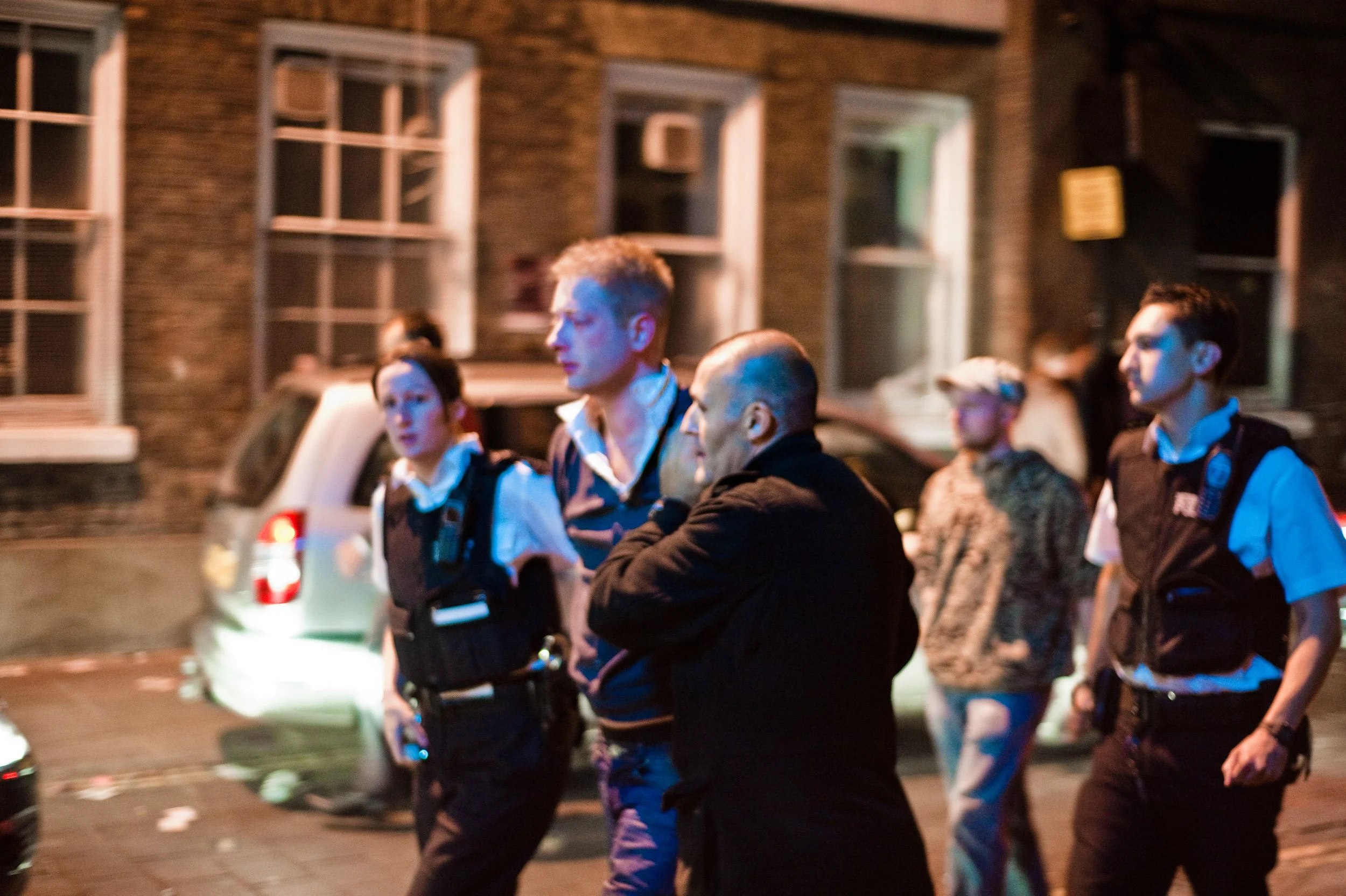 Nighttime scene on a city street with police officers and civilians. Several police officers in uniform are present, some with body cameras. A woman in a patterned jacket and a man in a black jacket are also visible.