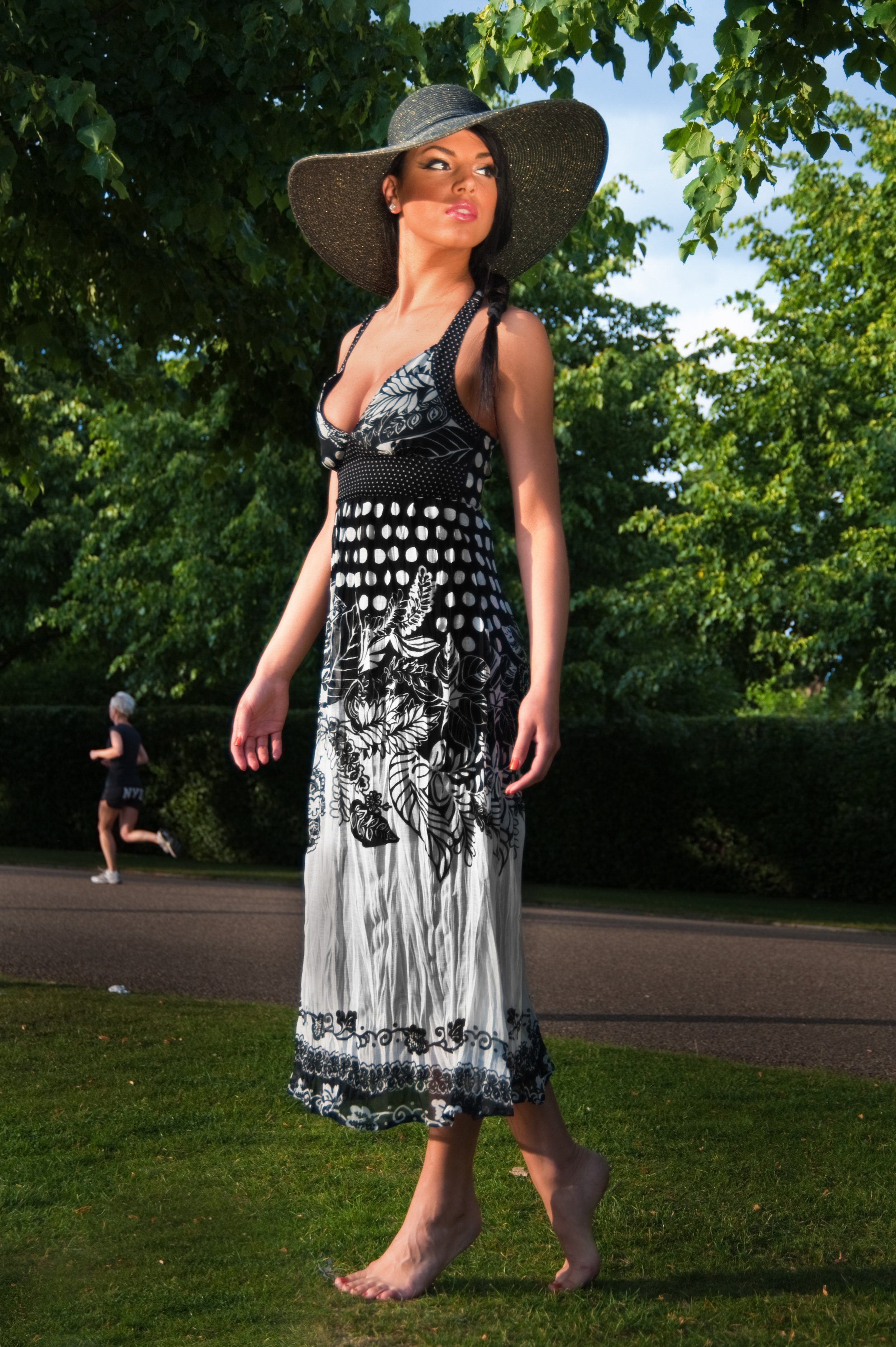 A woman in a black and white dress and large sun hat standing on grass in a park, with a jogging runner in the background.