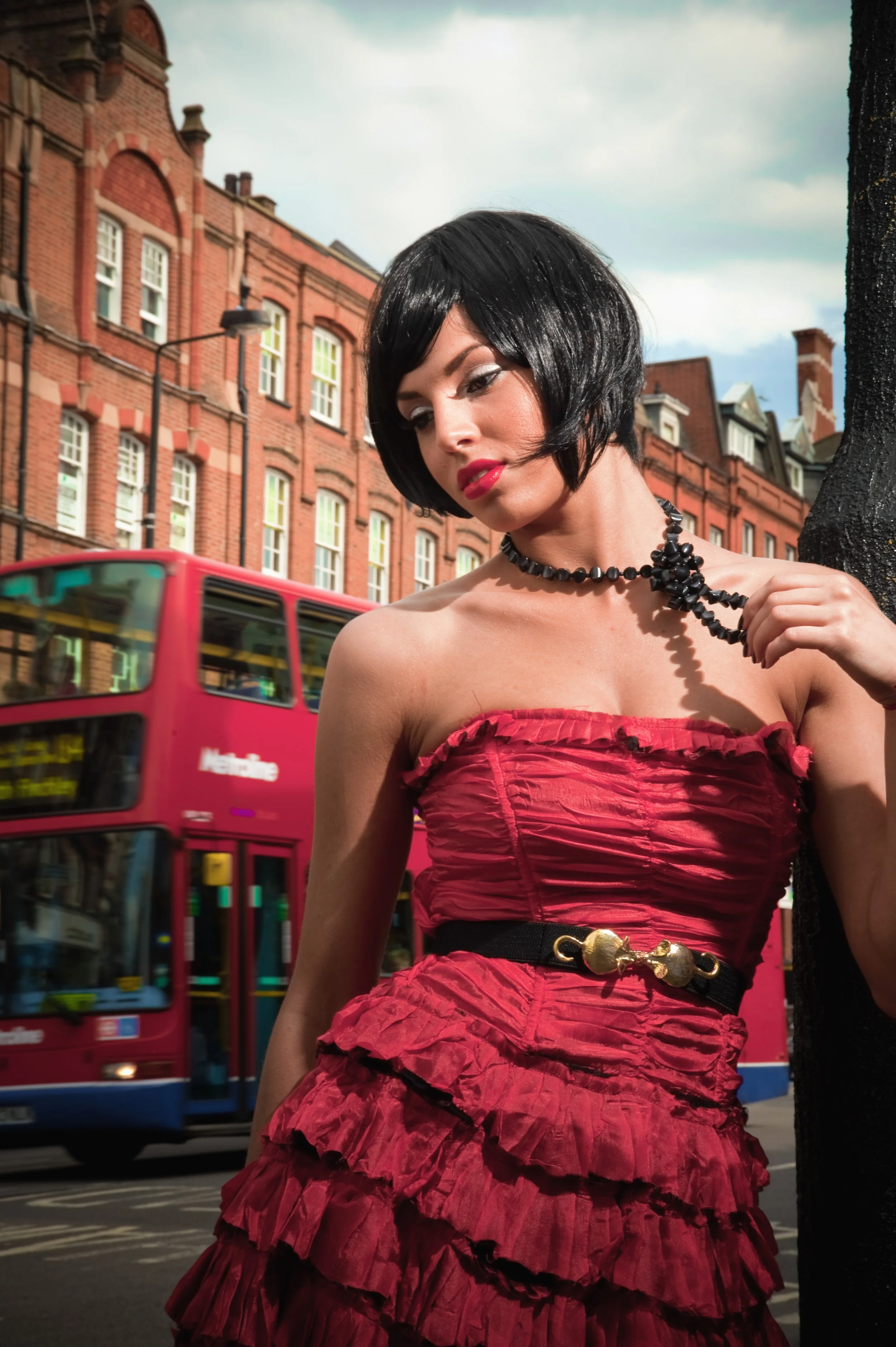 A woman with short black hair wearing a strapless red dress with ruffles, black jewelry, and a belt with a gold clasp, stands on a city street next to a black pole, with a red bus and brick buildings in the background.