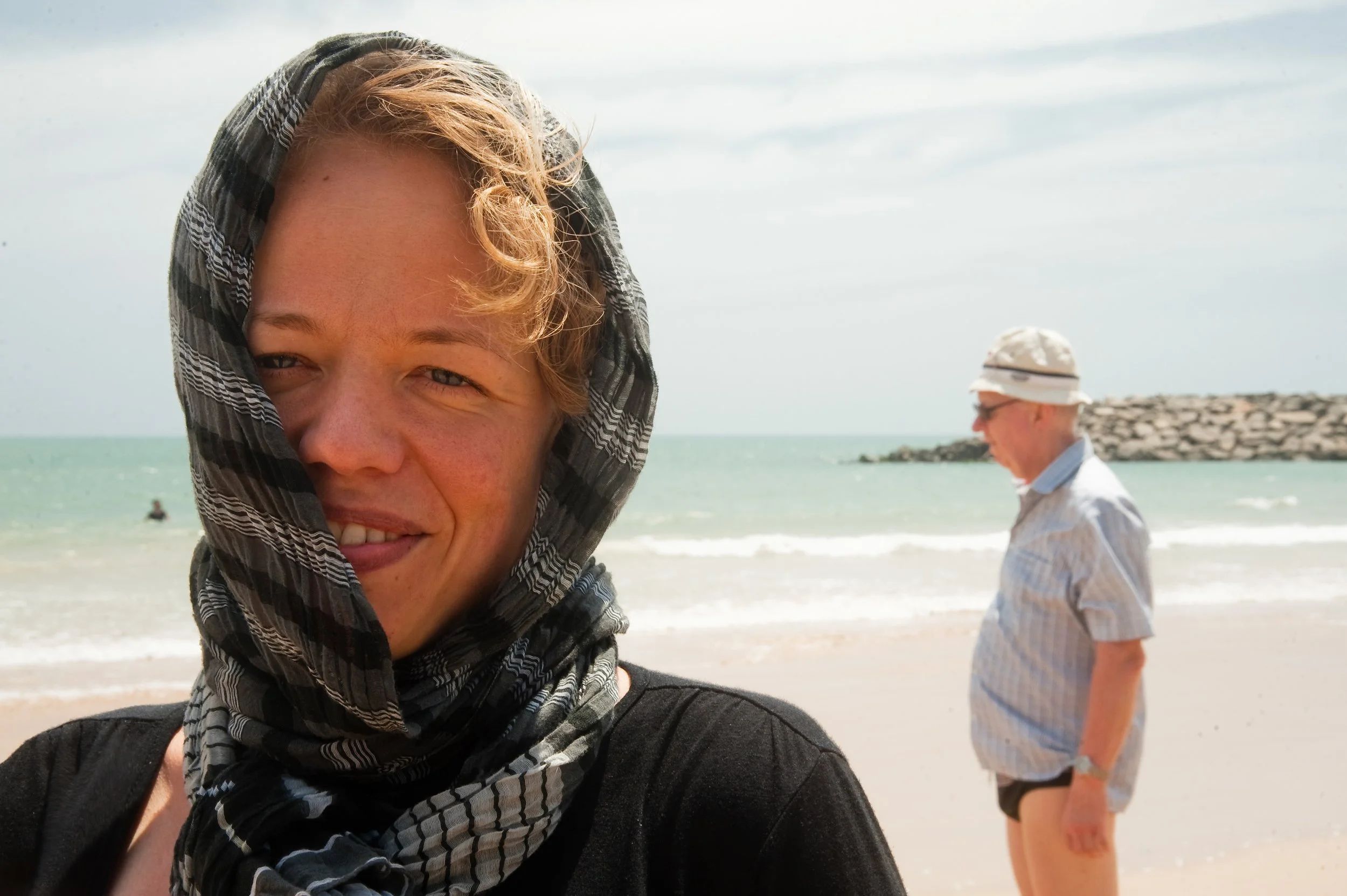 Close-up of a young woman smiling at the beach with a scarf on her head, with an elderly man walking in the background near the shoreline.