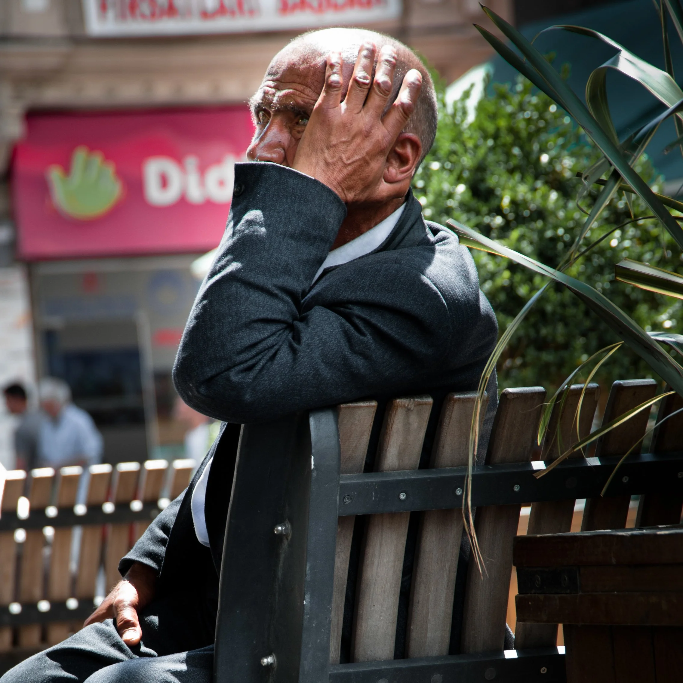 A man sitting on a park bench holding his head with his hand, looking distressed, with blurred background including a pink sign and people walking by.