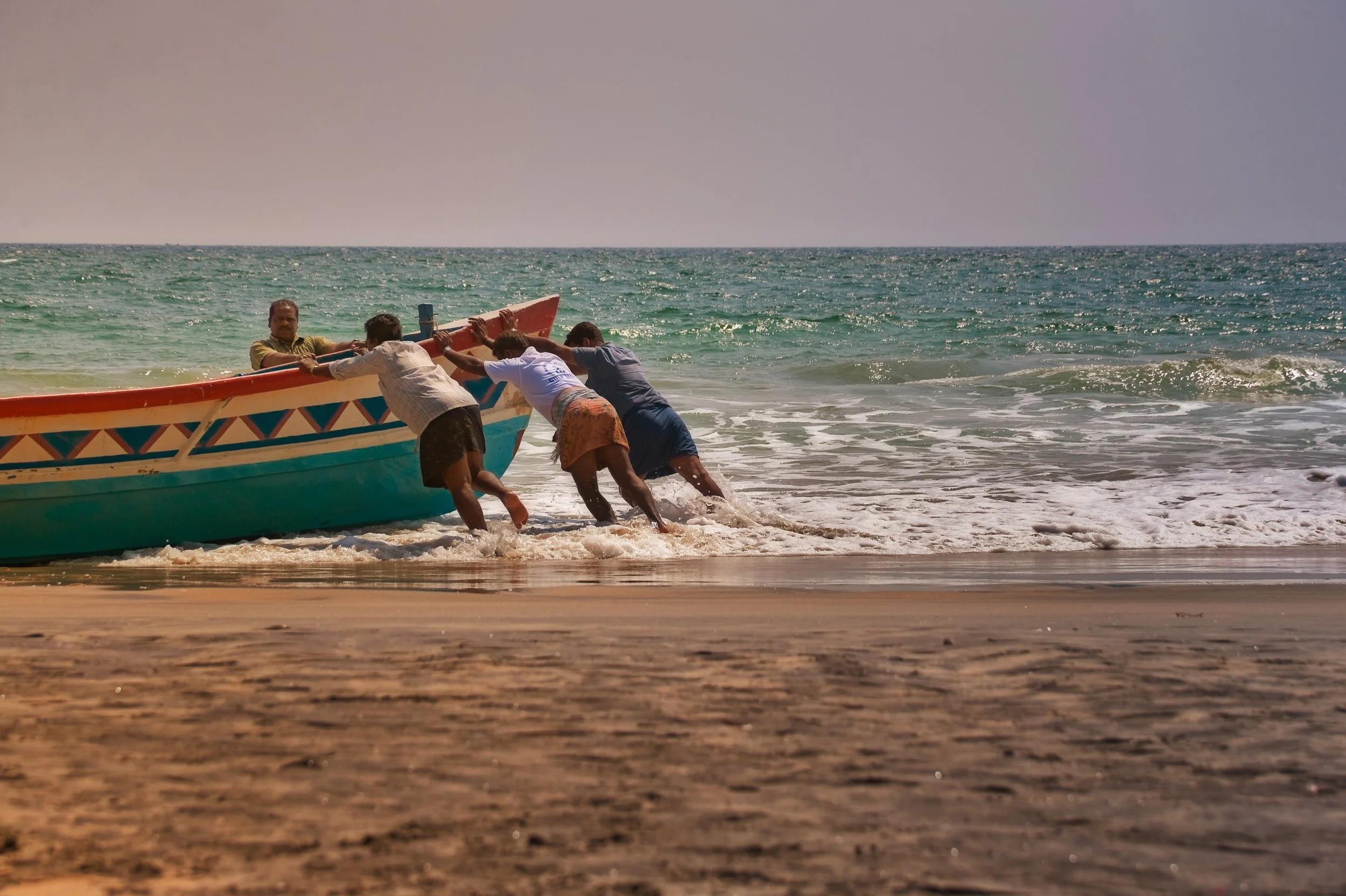Four men push a colorful boat into the ocean on a sandy beach near gentle waves under a clear sky.