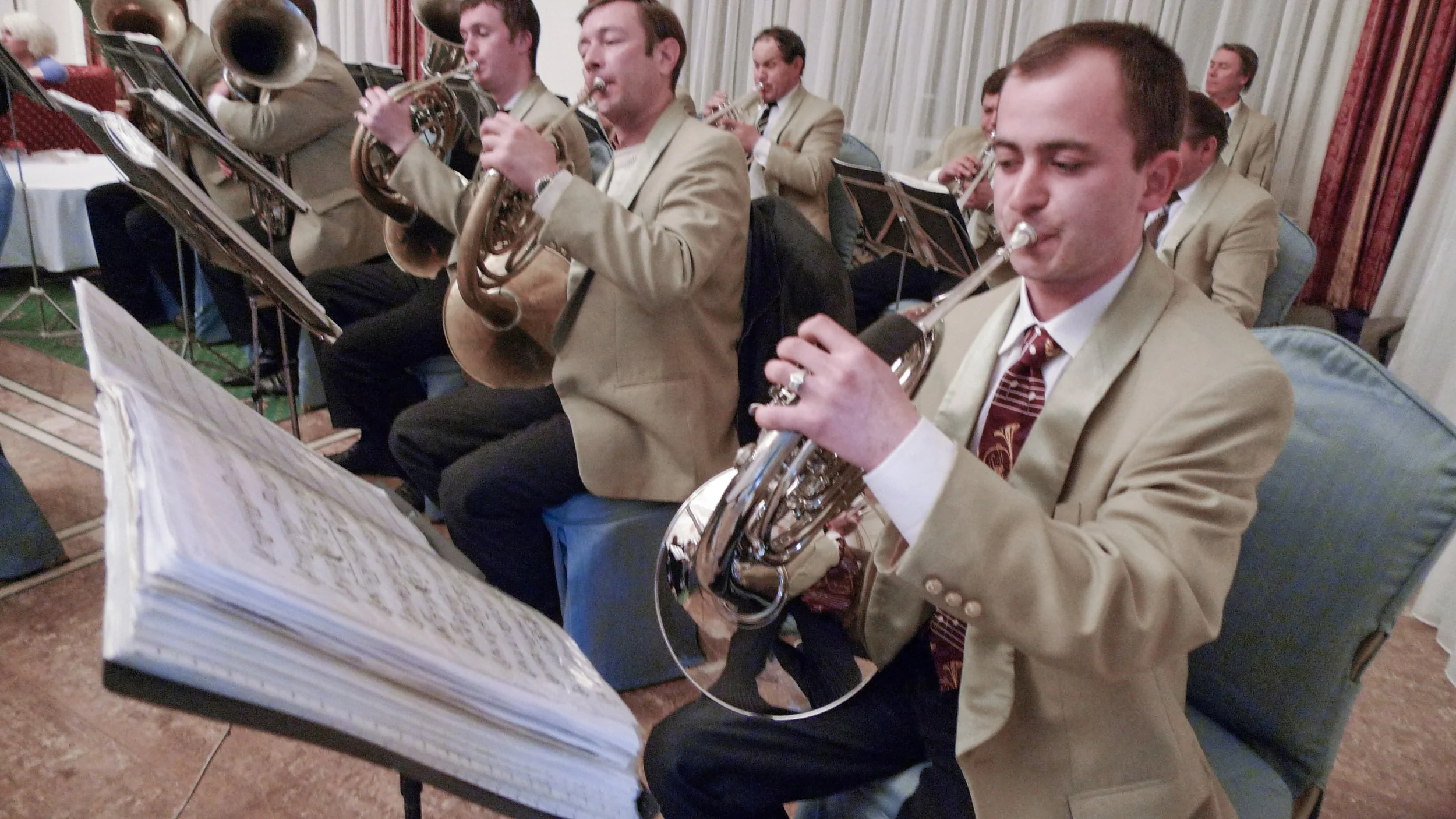 Musicians playing brass instruments in a band at a formal event, dressed in beige suits and seated in a decorated room with curtains.