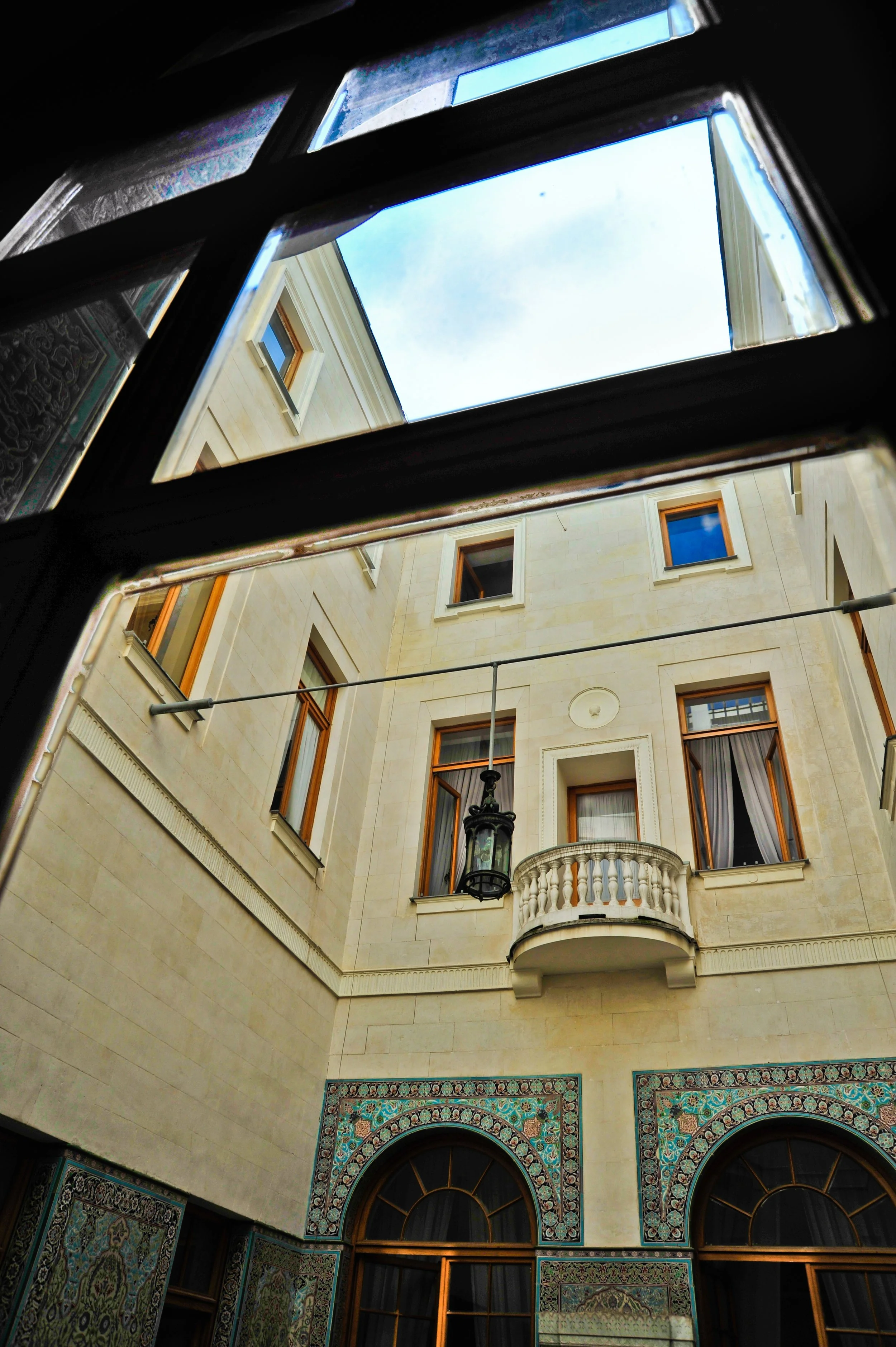 Looking up at a courtyard from a window with decorative tiles, showing windows with open shutters, a balcony, a lantern, and a blue sky.
