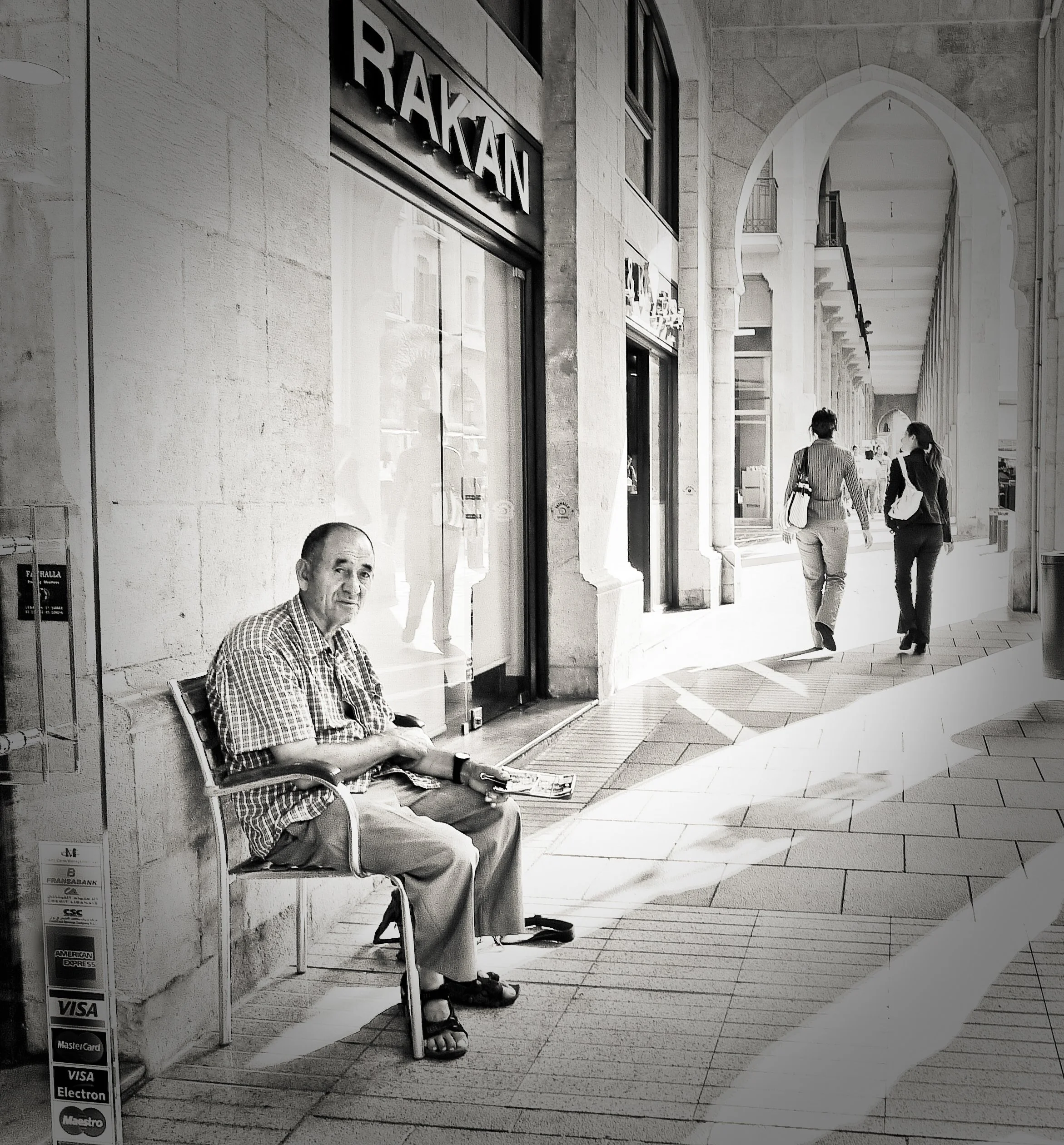 A man sitting on a bench outside a building with the sign 'RAKAM' above him, while two women walk along a covered arcade.