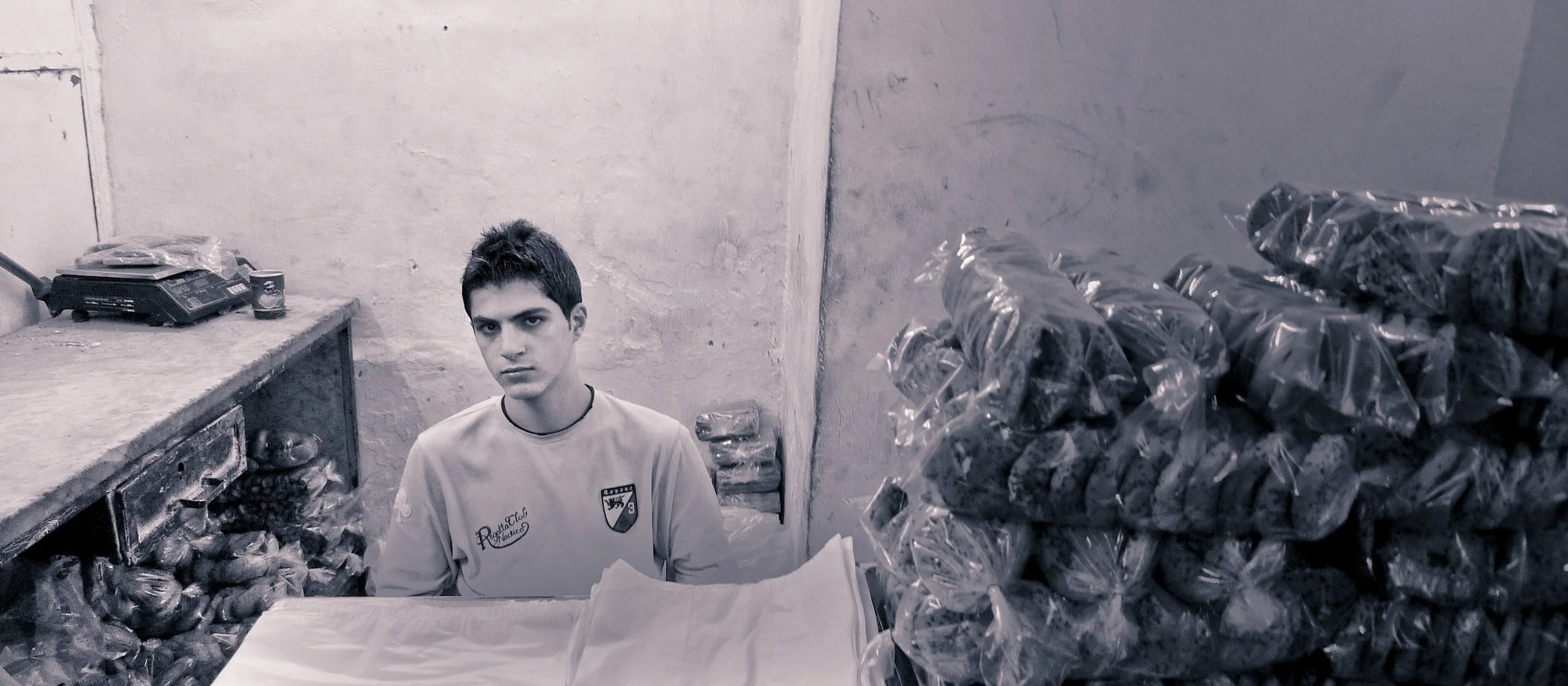 Young man sitting behind a counter surrounded by stacked plastic bags filled with dark objects in a bare room with unfinished walls.