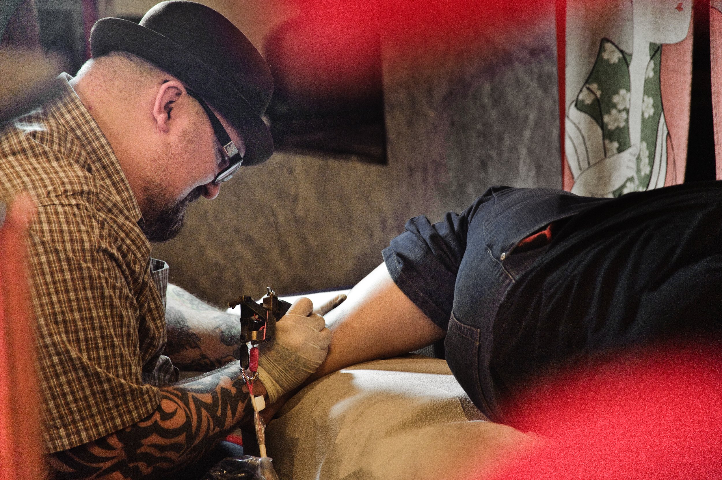 Tattooed man with a hat and glasses getting a tattoo on his upper arm by a tattoo artist wearing gloves.