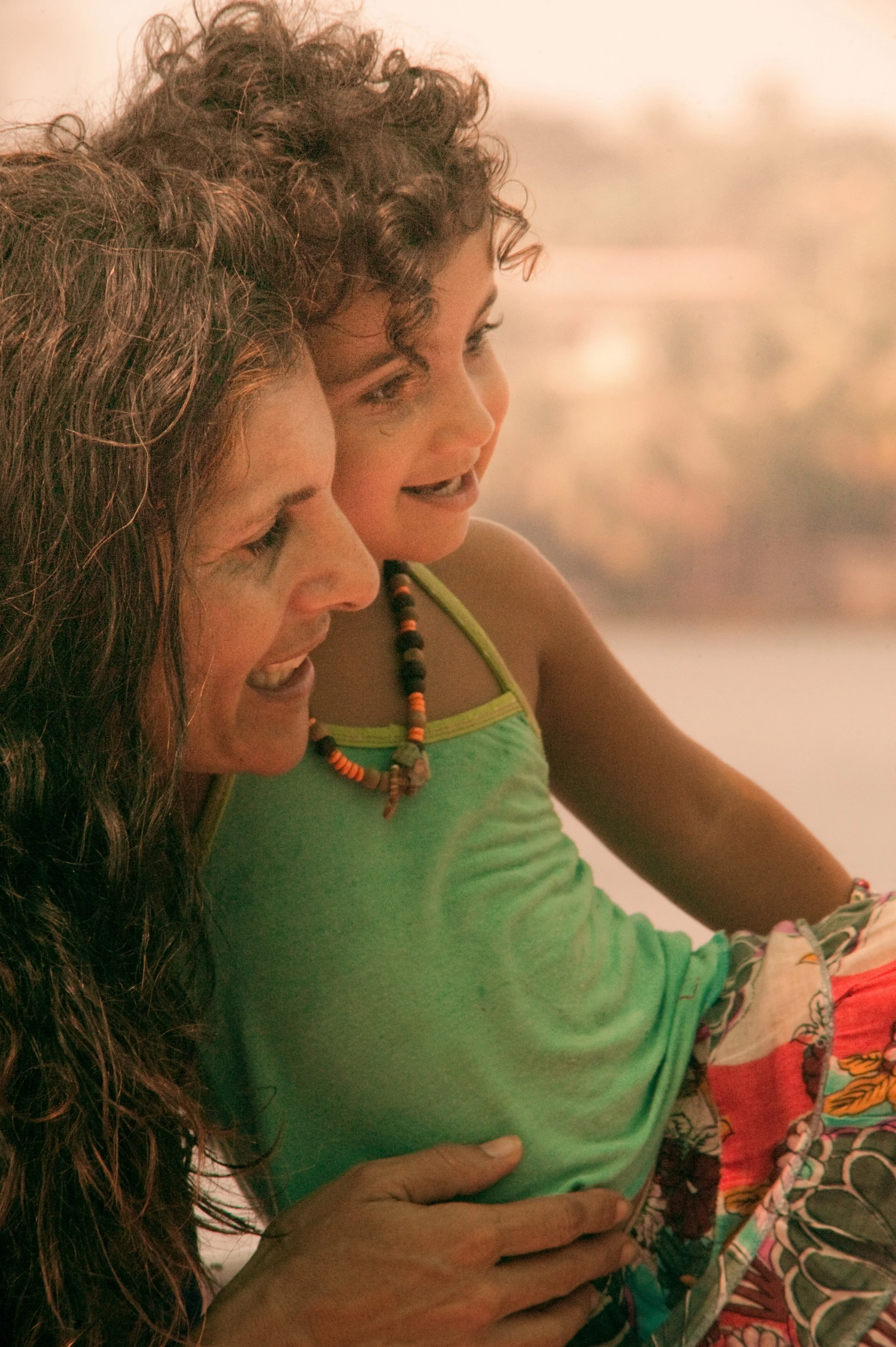 Two women smiling and looking at something, one with long wavy hair and the other with short curly hair, wearing colorful clothing and necklaces, with a soft focus background.