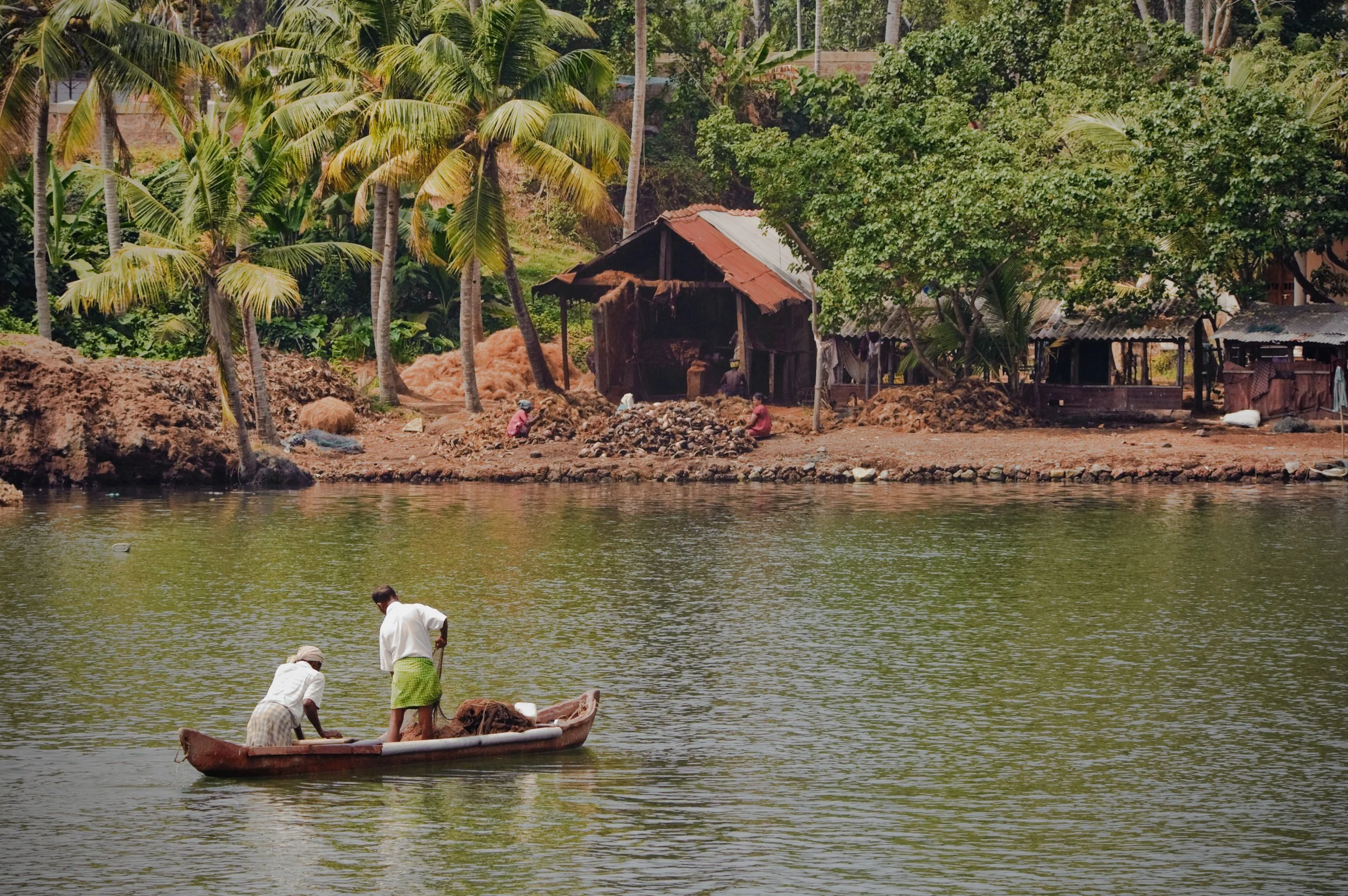 Two men in a small boat on a river, with a riverside village in the background, featuring trees, wooden huts, and people working near the shore.