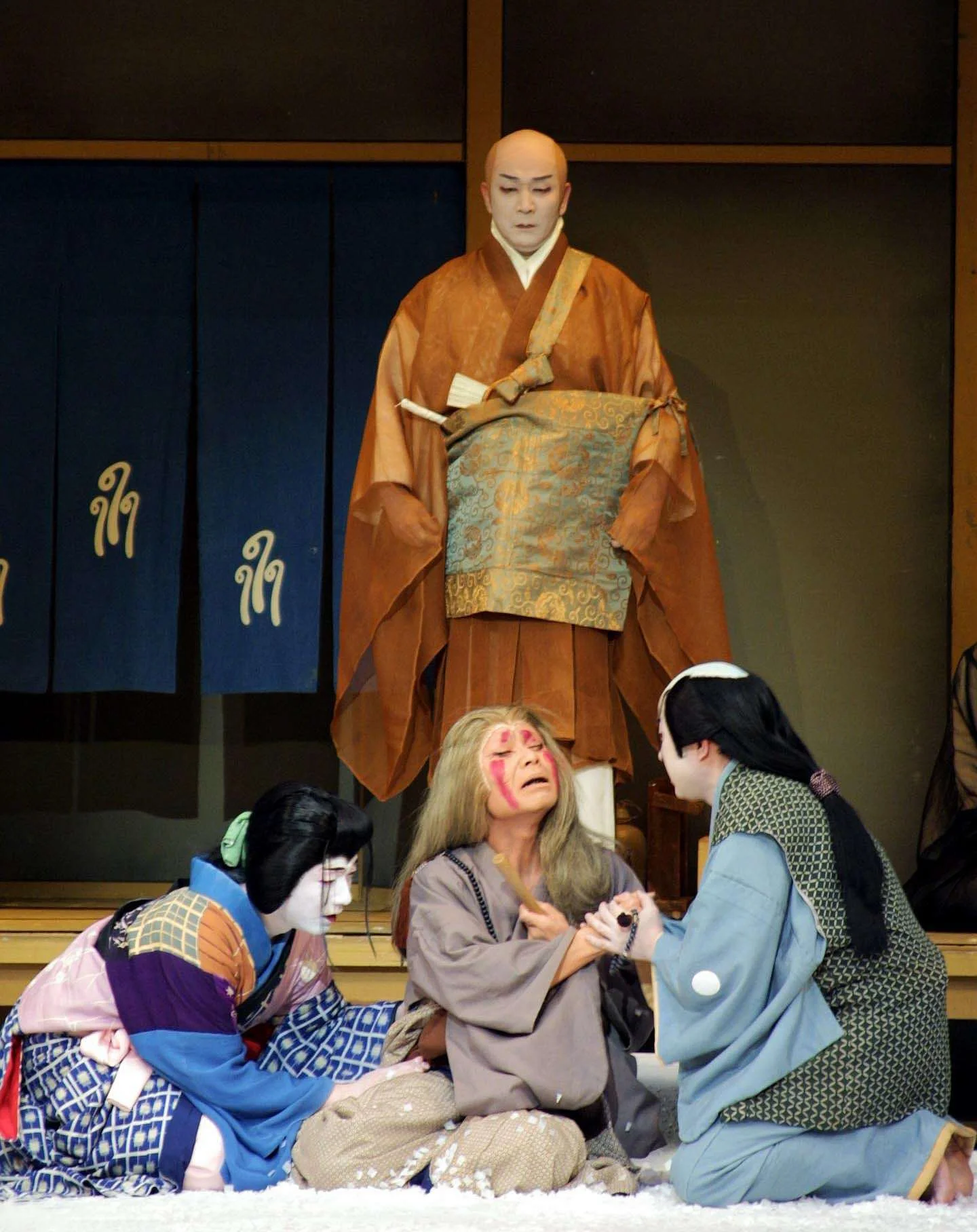 A traditional Japanese scene with actors in kimono costumes performing a play on stage. One actor is in the background in a large costume, while three actors in the foreground are kneeling and interacting.