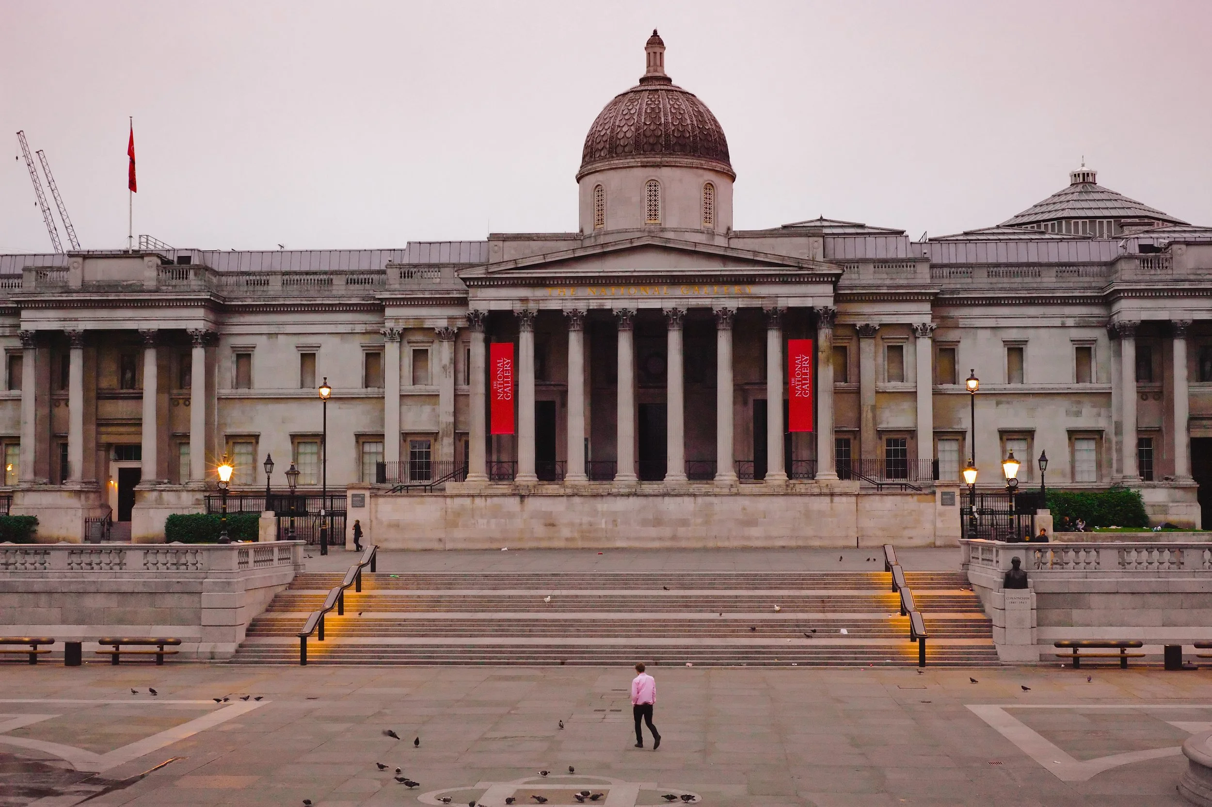 The National Gallery building with columns and a dome, stairs leading up to the entrance, and a person walking in front on a paved area with pigeons.