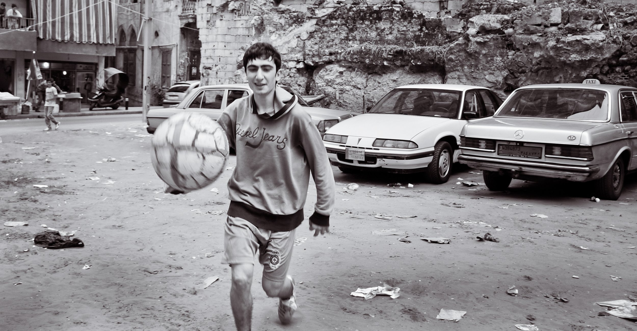 A young boy playing soccer on a dirt street with cars parked in the background, some pedestrians, and a rocky wall behind parked cars.