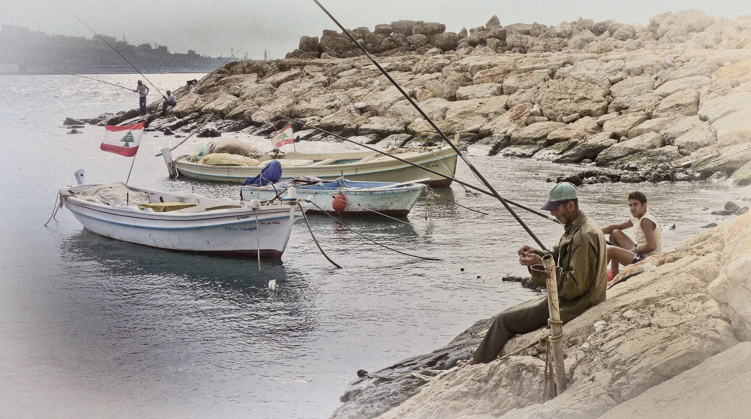 Two men fishing on a rocky shoreline near three boats with Lebanese flags, with another man sitting on the rocks.