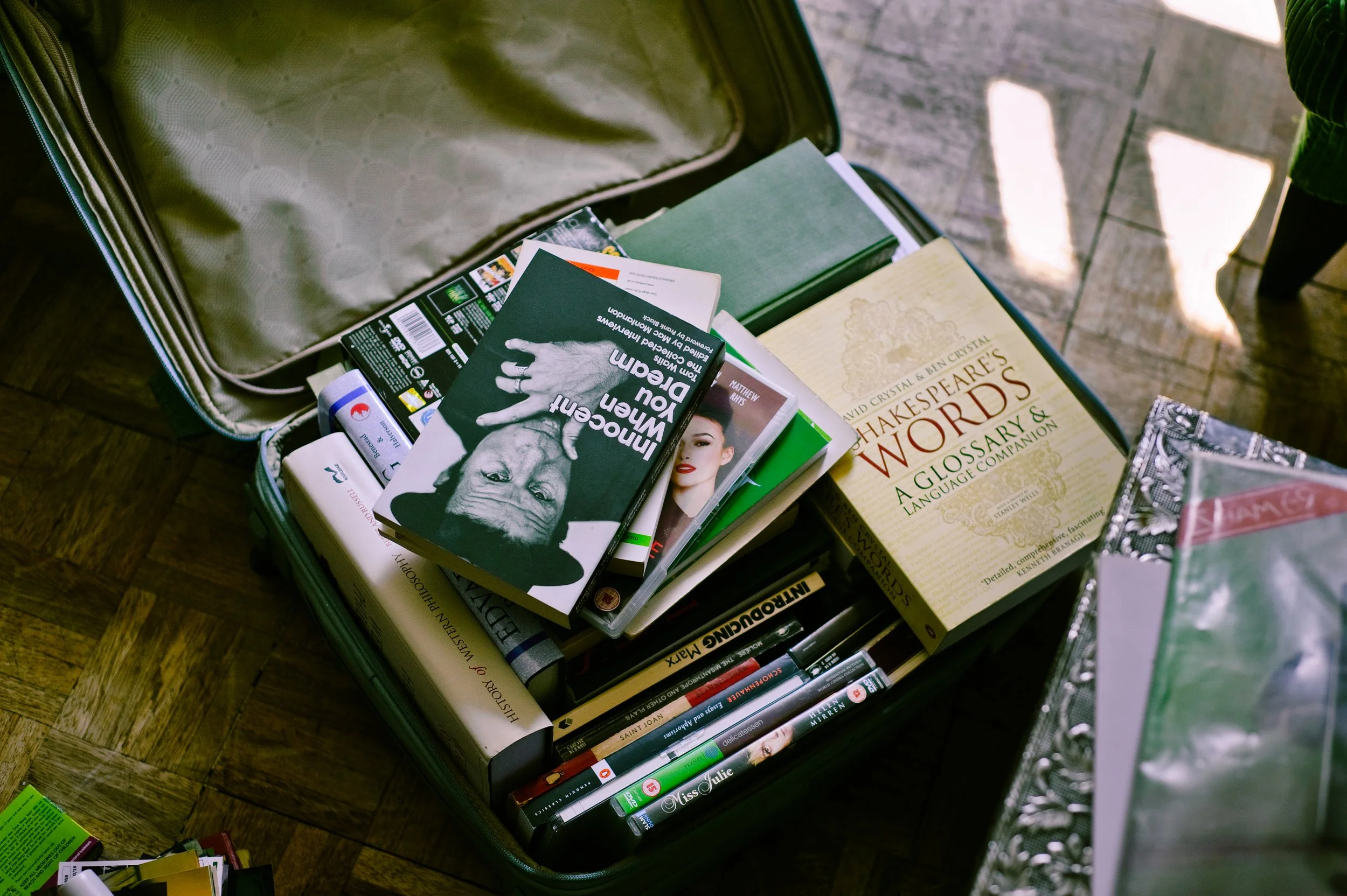 Open green suitcase filled with books and magazines, including a book titled 'Shakespeare's Words' and a book titled 'Imagine You Dream.'