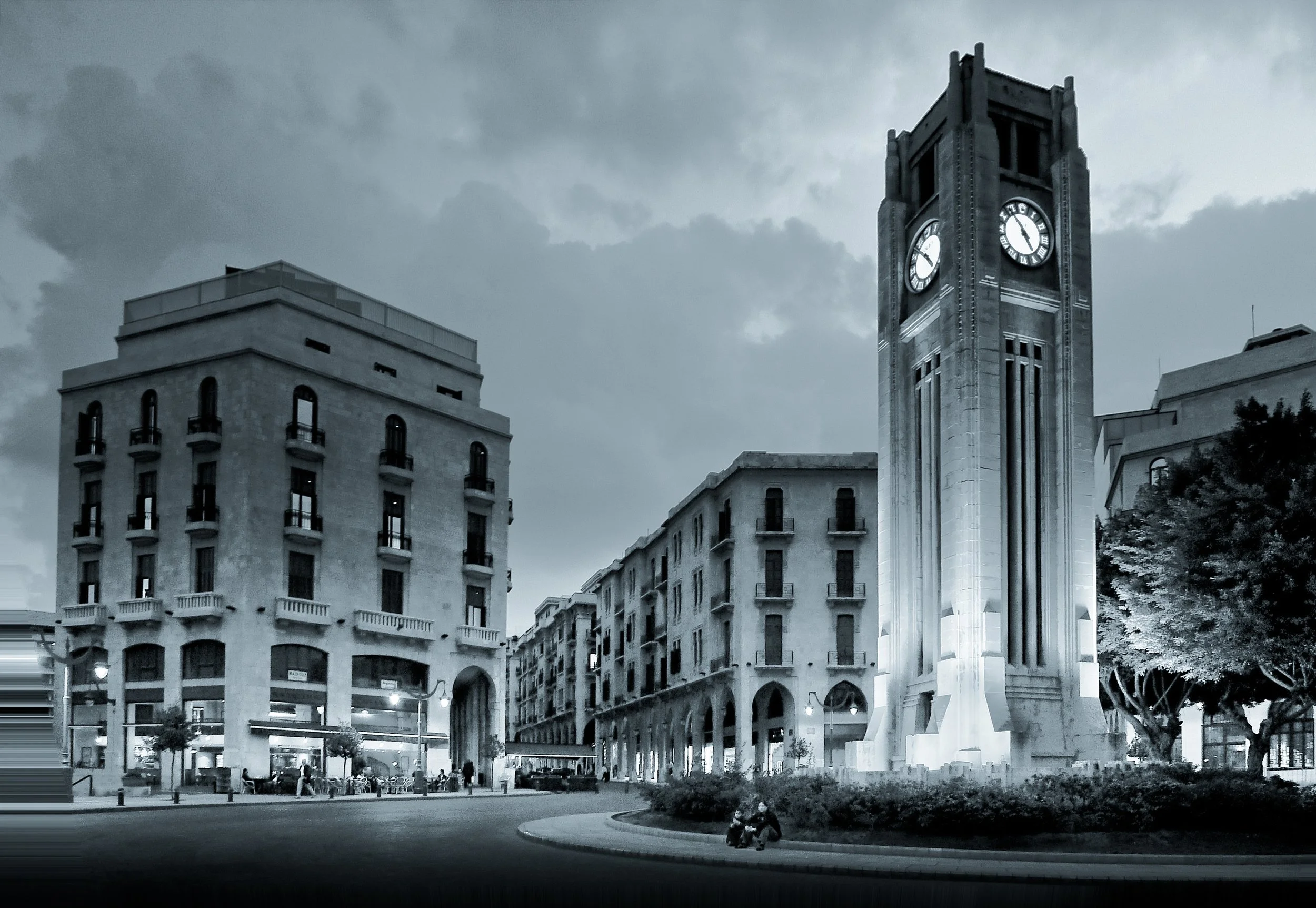 Nighttime cityscape with a tall clock tower illuminated, surrounded by historic buildings and trees, under cloudy sky.