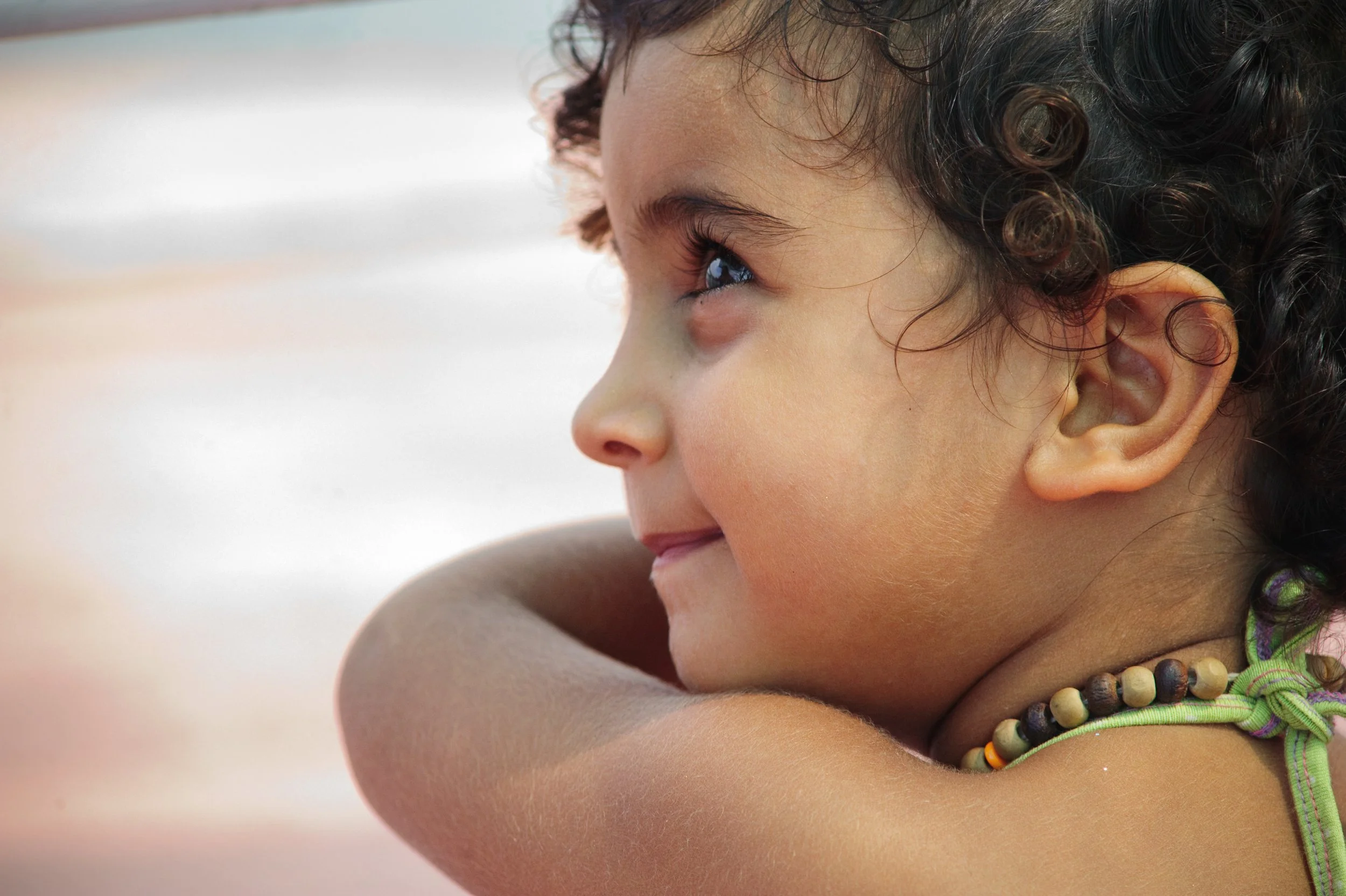 Close-up of a young girl with curly dark hair, wearing a beaded necklace and a green strap top, resting her chin on her arms and gazing into the distance with a smile.