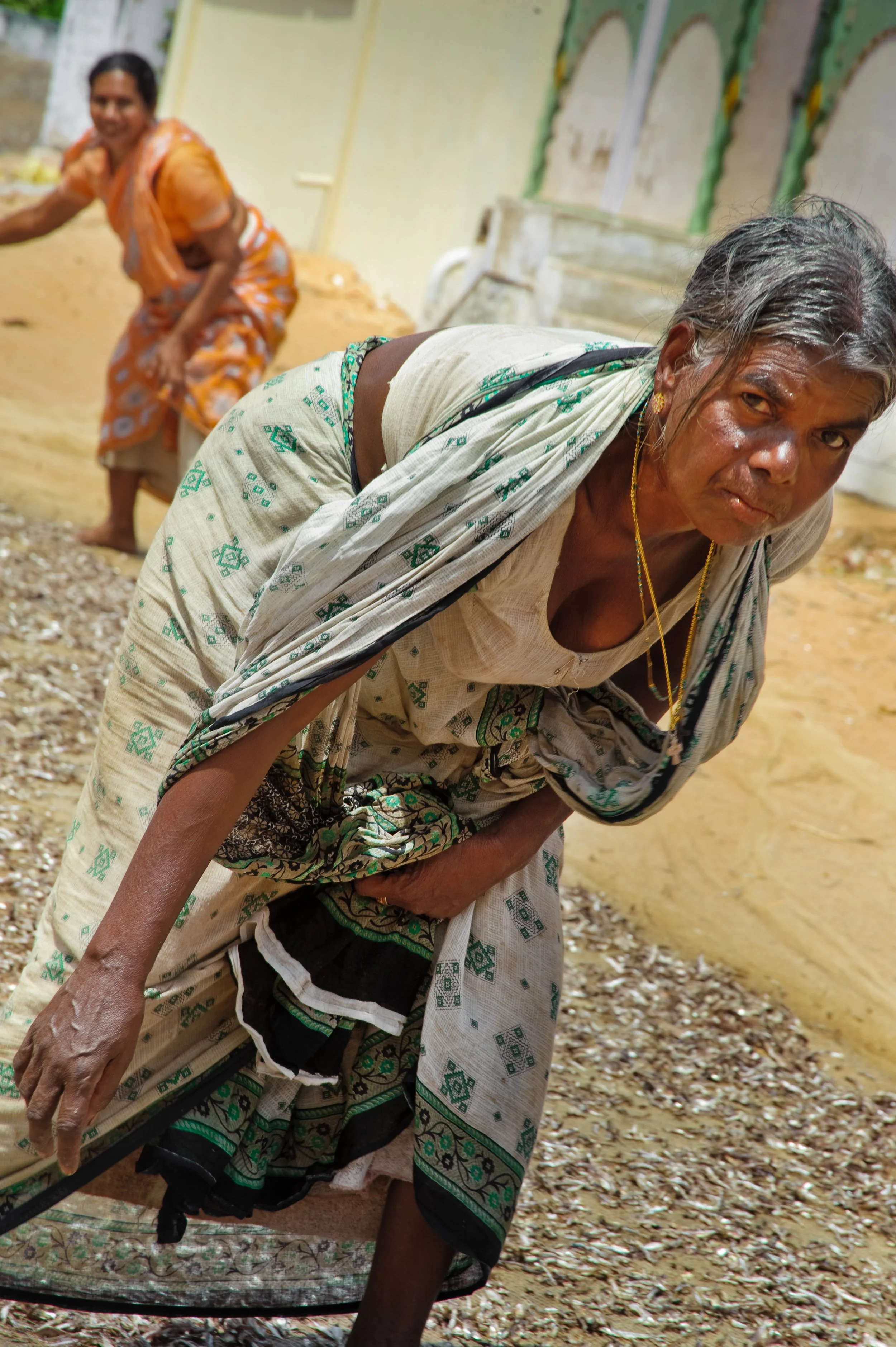 An elderly woman bends over holding a stick, wearing traditional patterned clothing, with a smiling woman in the background.