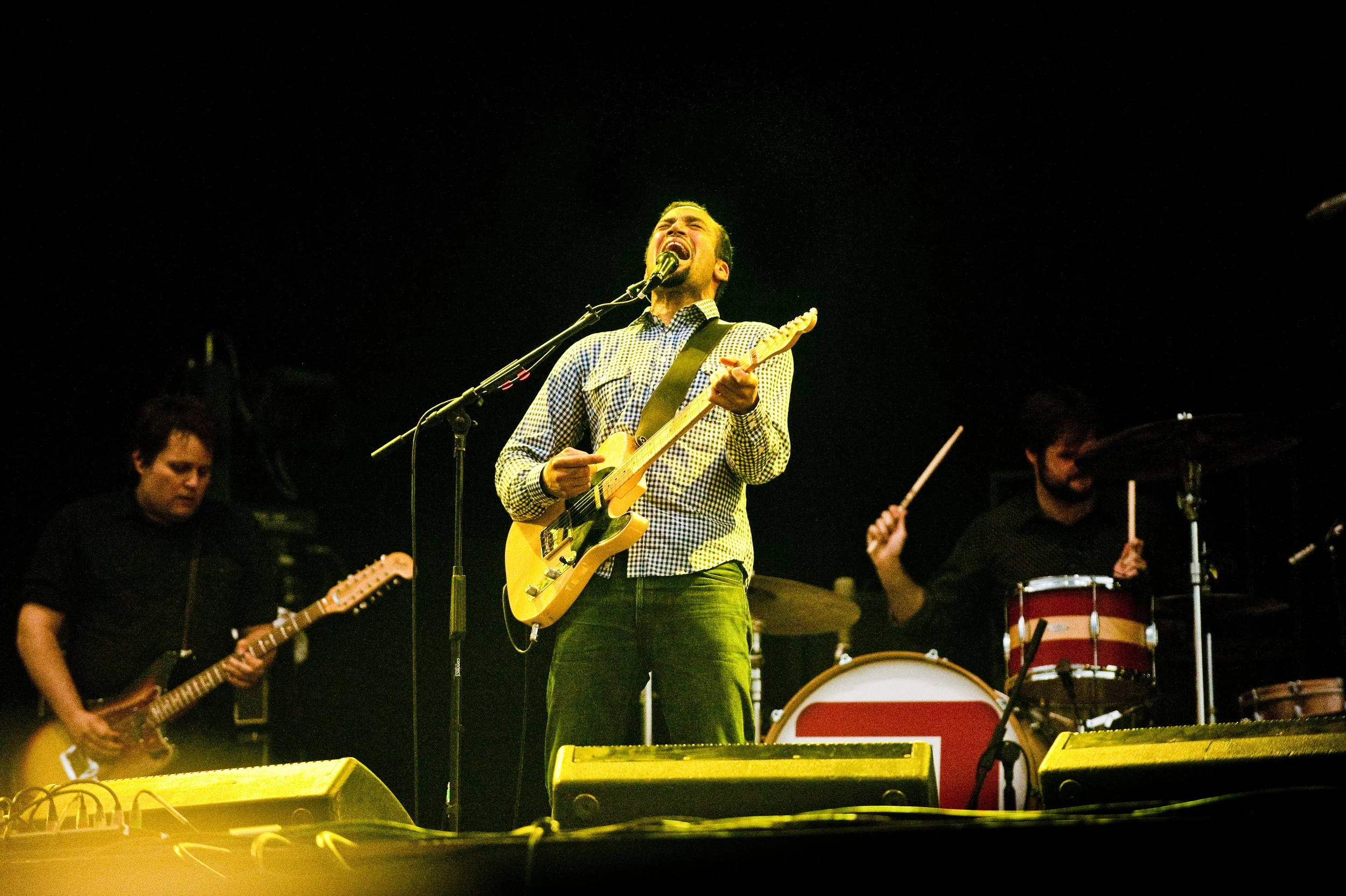 A band performs on stage, with a lead singer and guitarist in the center singing into a microphone, flanked by a guitarist on the left and a drummer on the right.