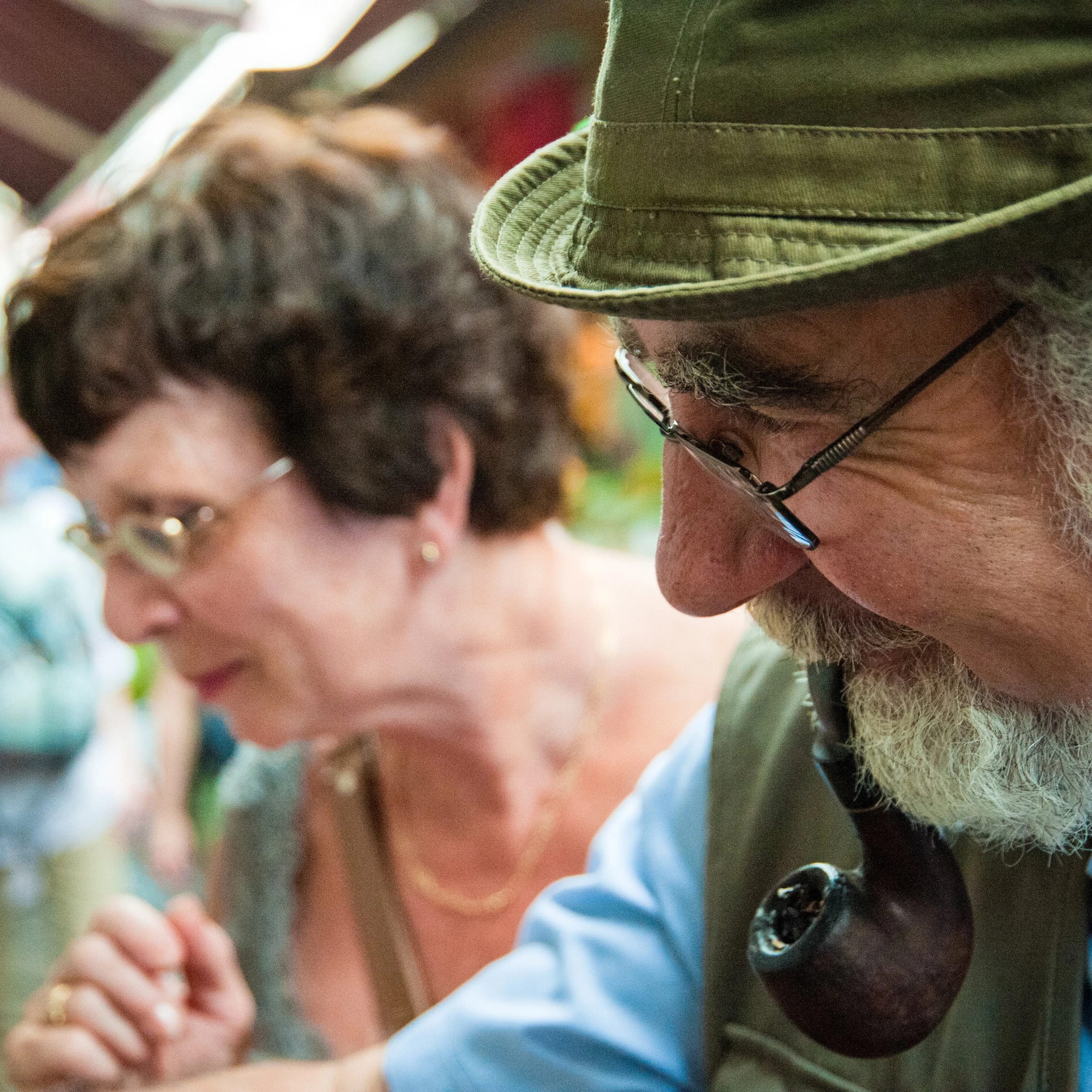 Close-up of a man with glasses, a beard, and wearing a green hat, looking downward. A woman with glasses and short dark hair is blurred in the background.