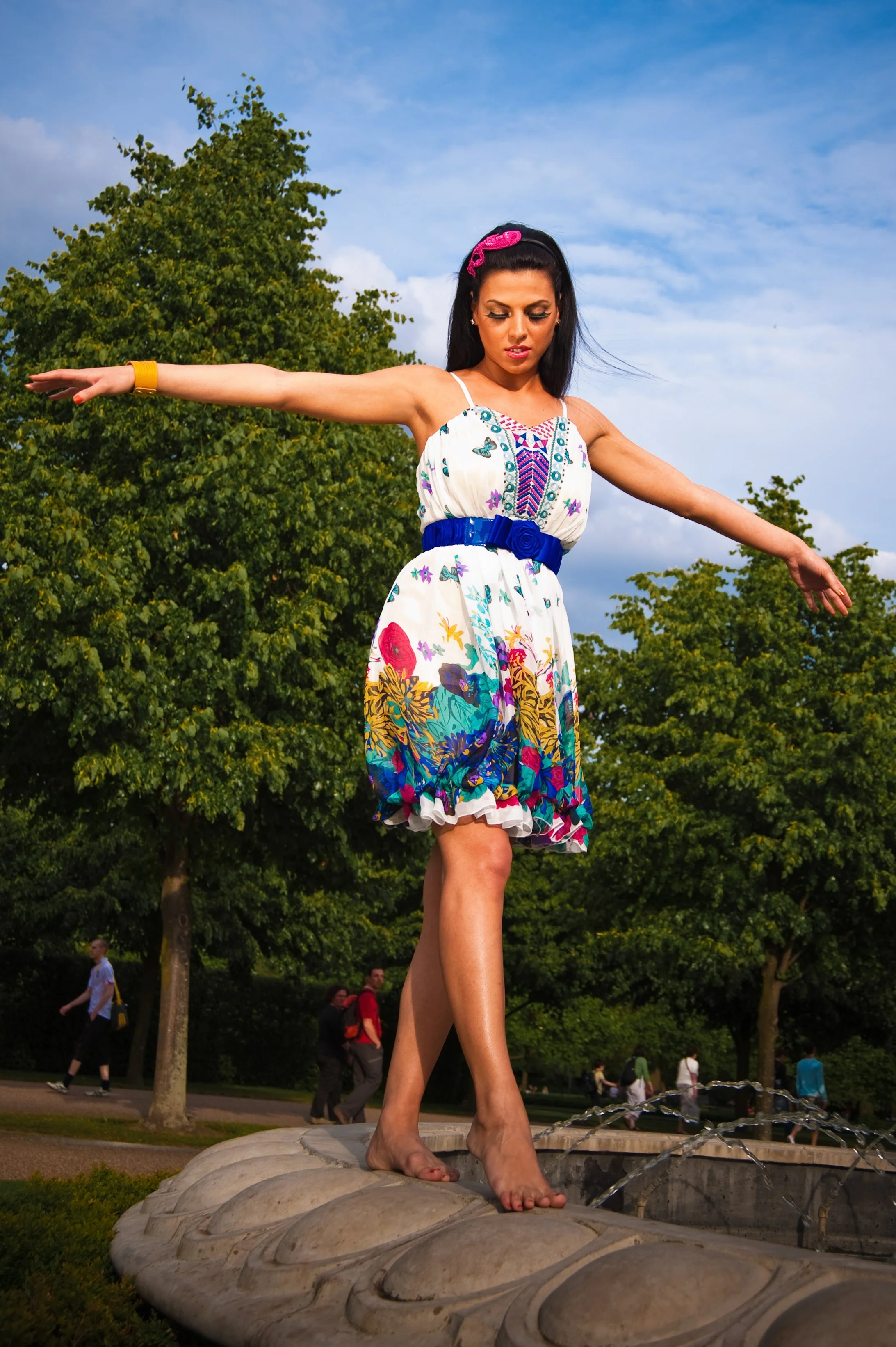 A woman standing on a fountain during daytime, wearing a colorful dress and a pink headband, with arms extended and looking downward.