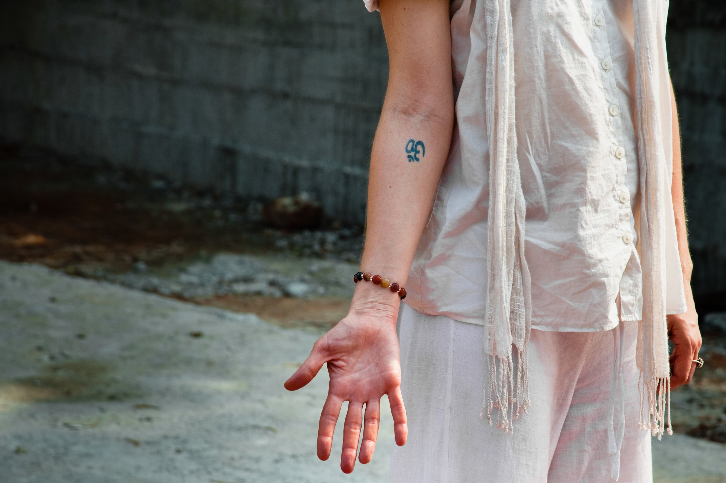 Close-up of a person's arm and hand, wearing a beaded bracelet, a blue tattoo, and a ring, with clothing in light colors, standing outdoors on a concrete surface with a blurred background.