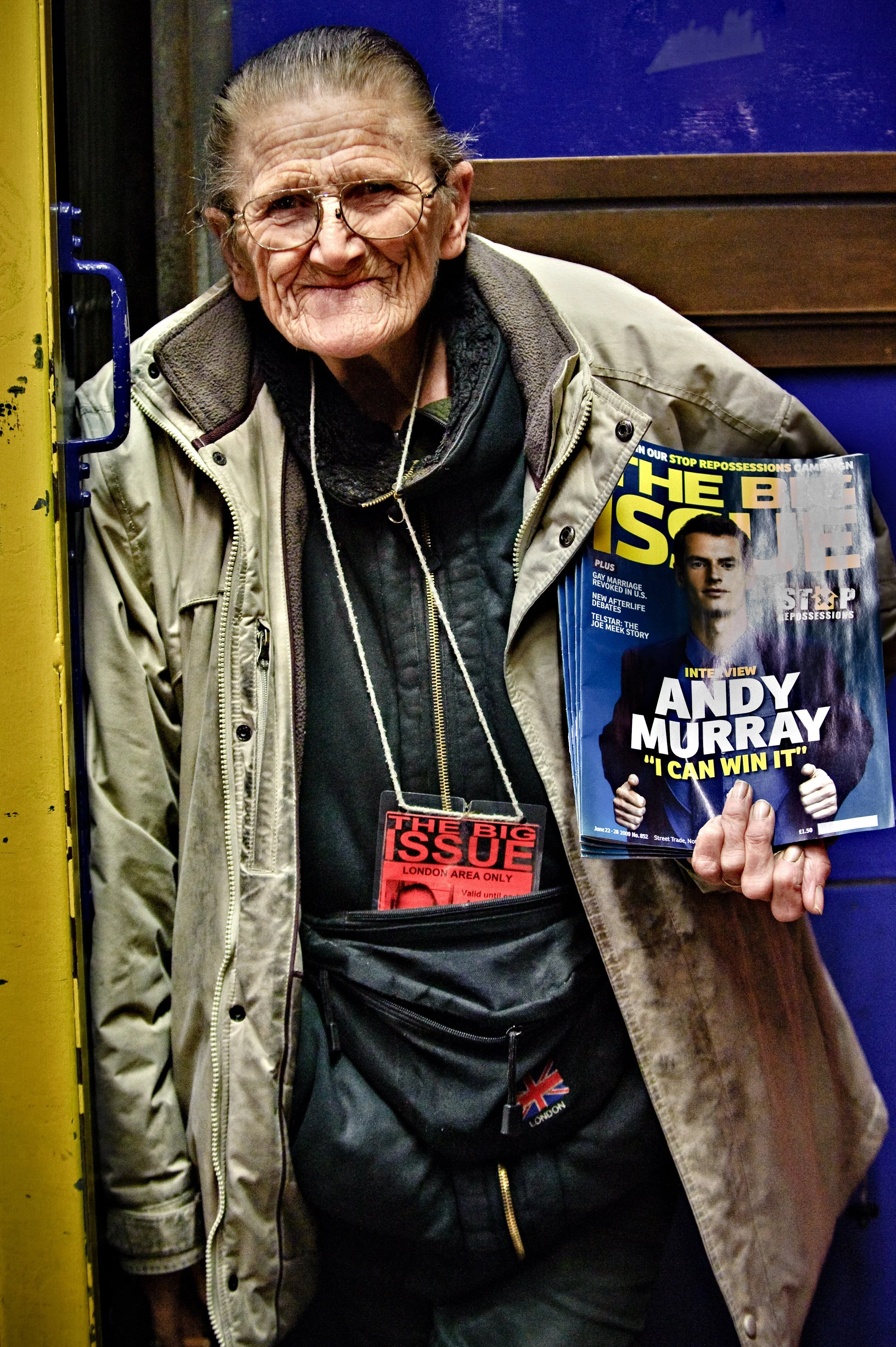 An older woman with glasses holding a magazine that features an interview with Andy Murray, titled "I CAN WIN IT." She is standing in front of a yellow and blue background, wearing a beige jacket and a black waist pouch.