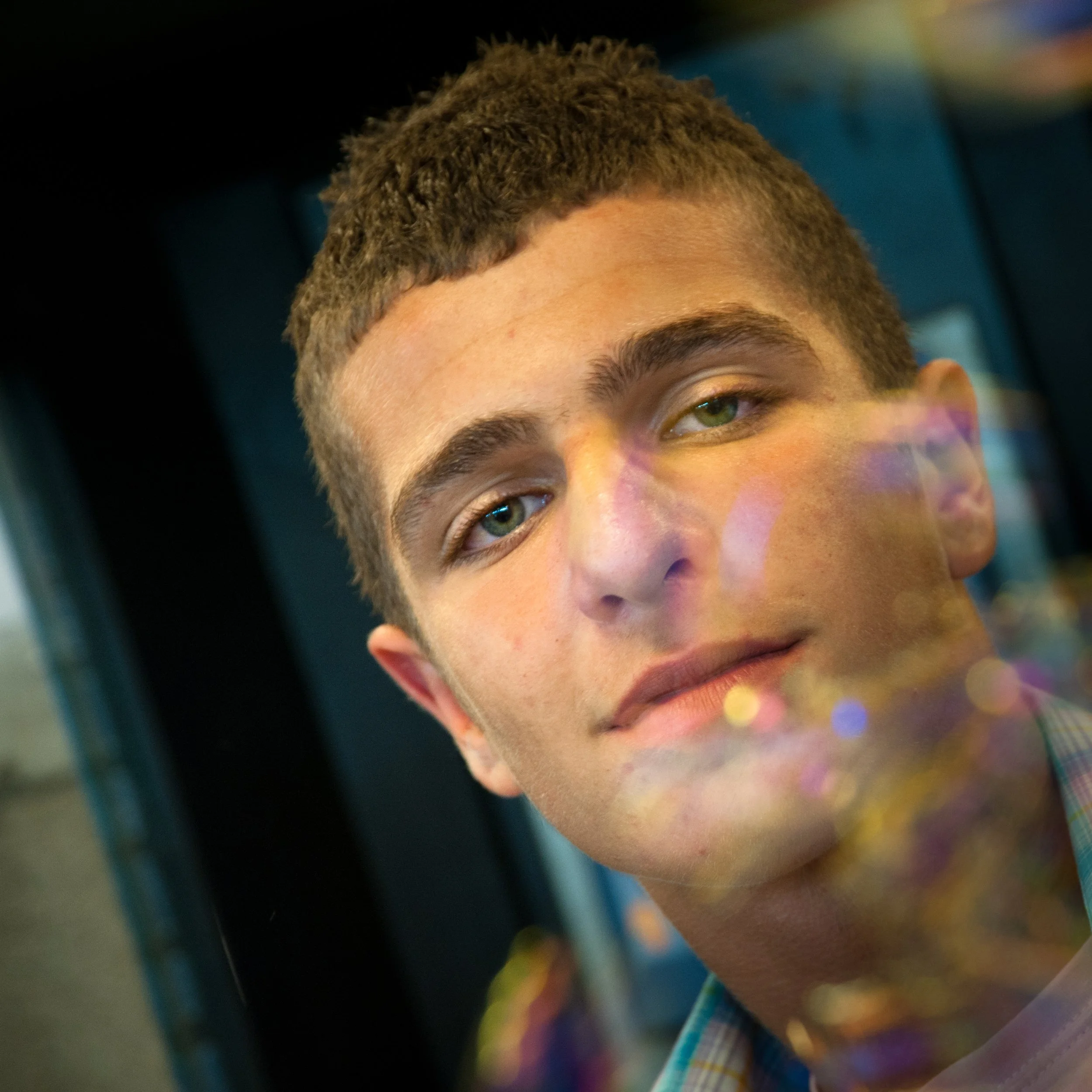 Close-up of a young man with short, curly brown hair, light skin, and green eyes, looking at the camera with a slight smile.
