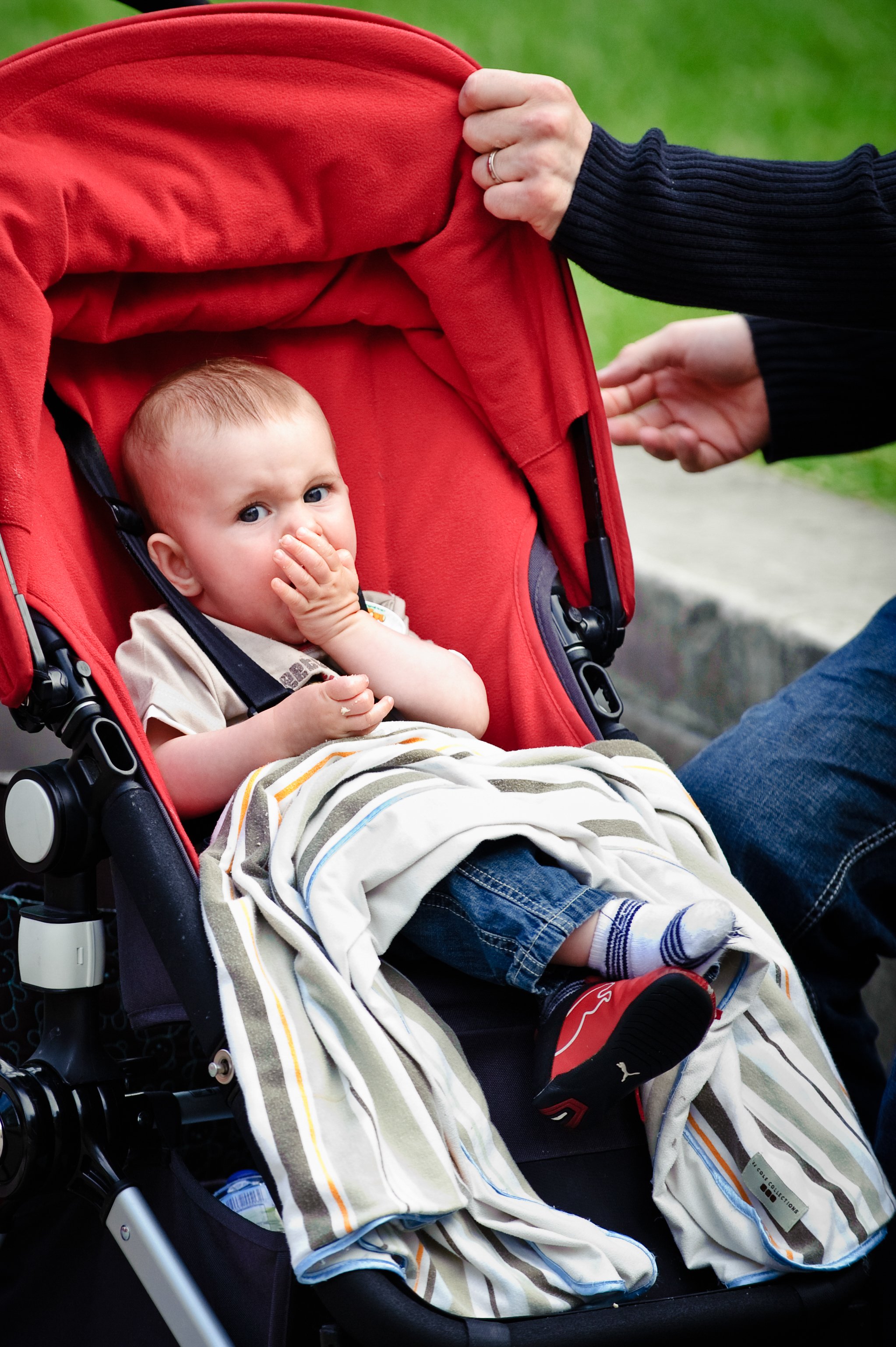A young child with light hair sitting in a red stroller, wearing a beige shirt, blue jeans, and striped socks, covering his mouth with one hand while looking at the camera. An adult in a black sweater is adjusting the stroller handle outside on a gra