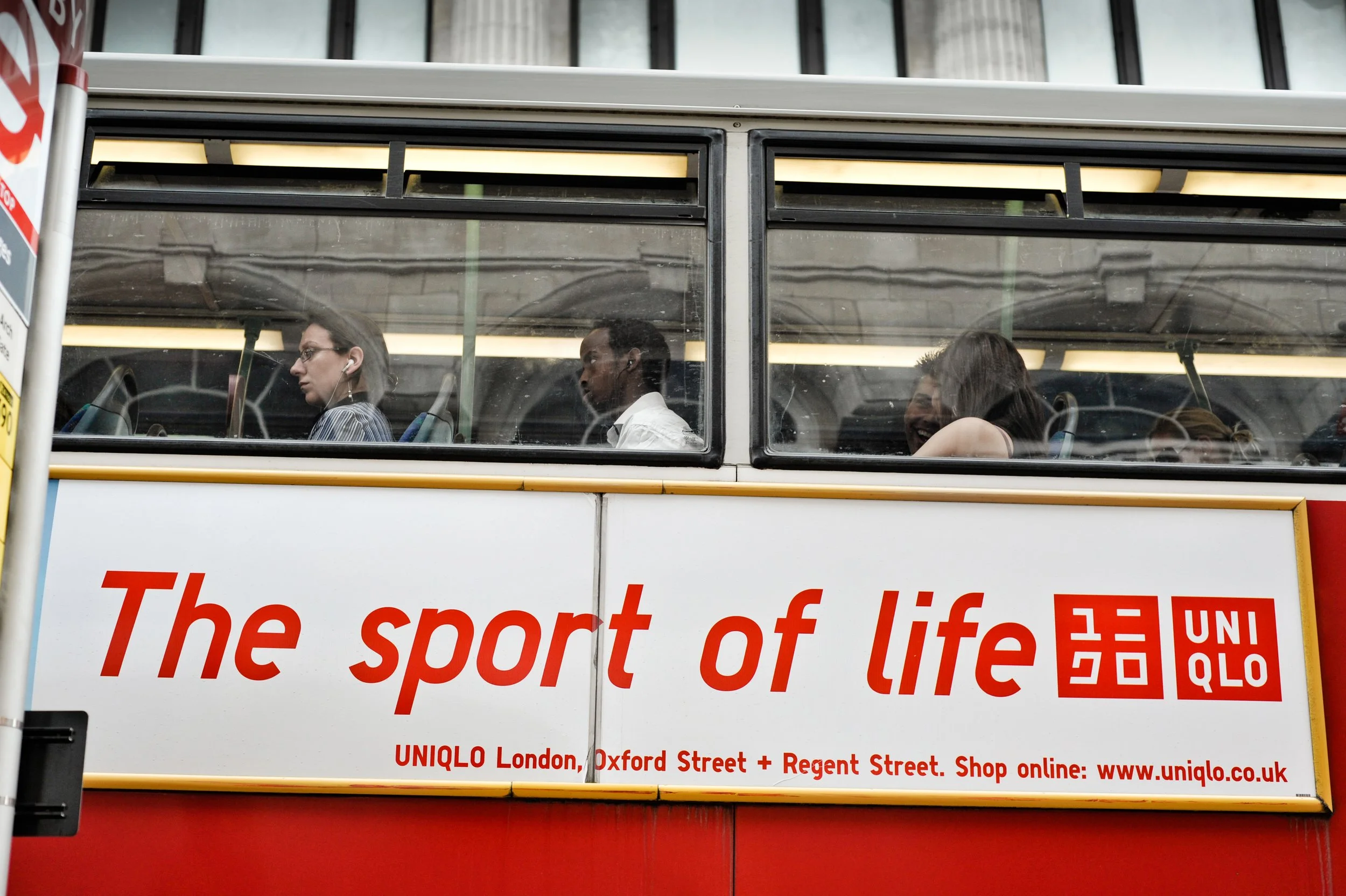 A double-decker bus with an advertisement for UNIQLO, displaying the slogan "The sport of life." There are four visible passengers inside, including two women and a man, some of whom are seated and some looking forward.