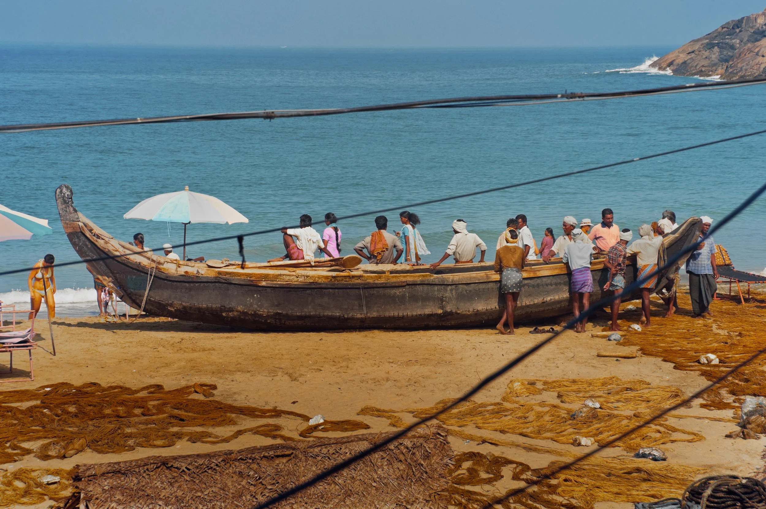 A group of people working on a wooden boat by the beach, with the ocean and rocky cliffs in the background, and fishing nets laid out on the sand in the foreground.