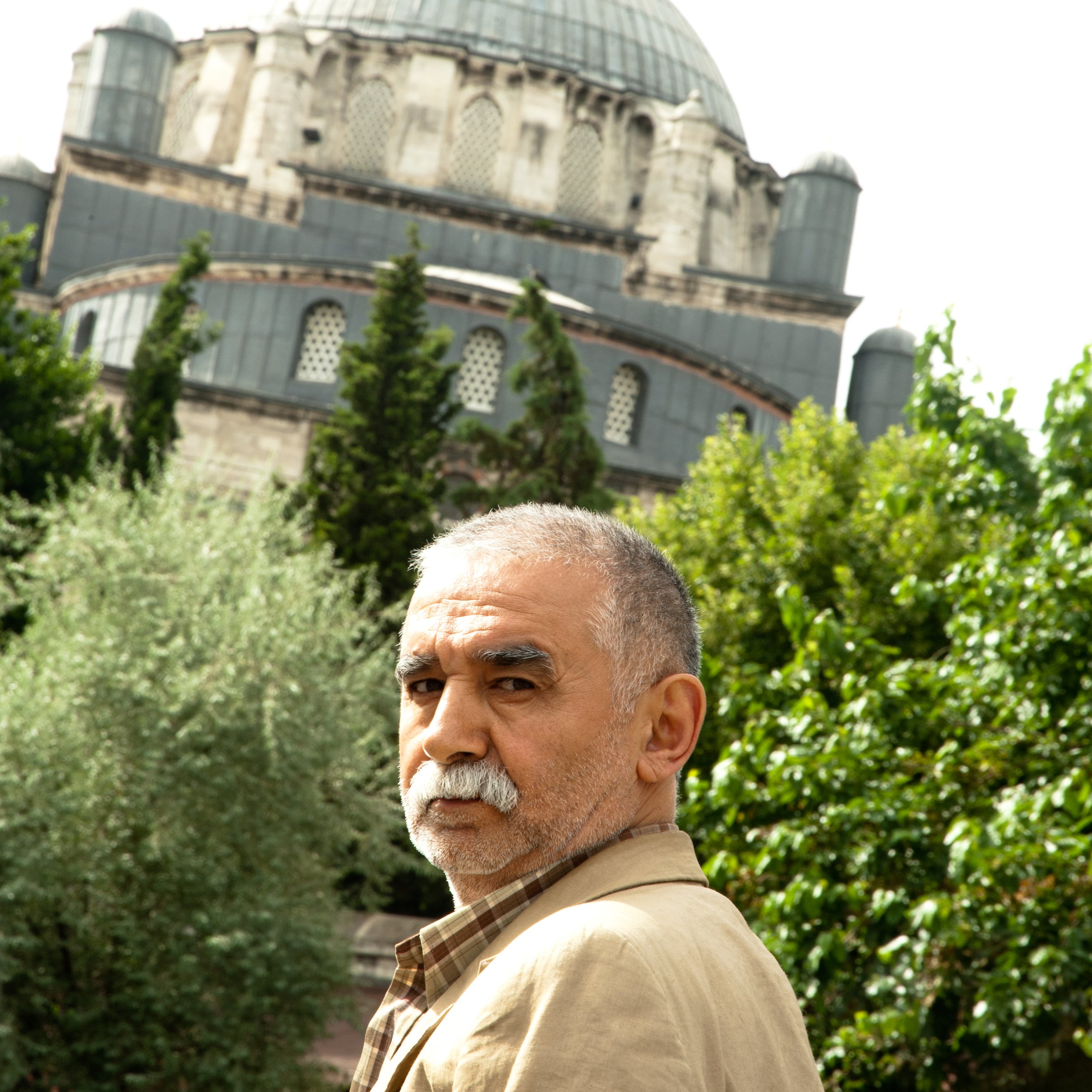 Older man with gray hair and mustache wearing a tan jacket, standing outdoors with green trees and a historic building in the background.
