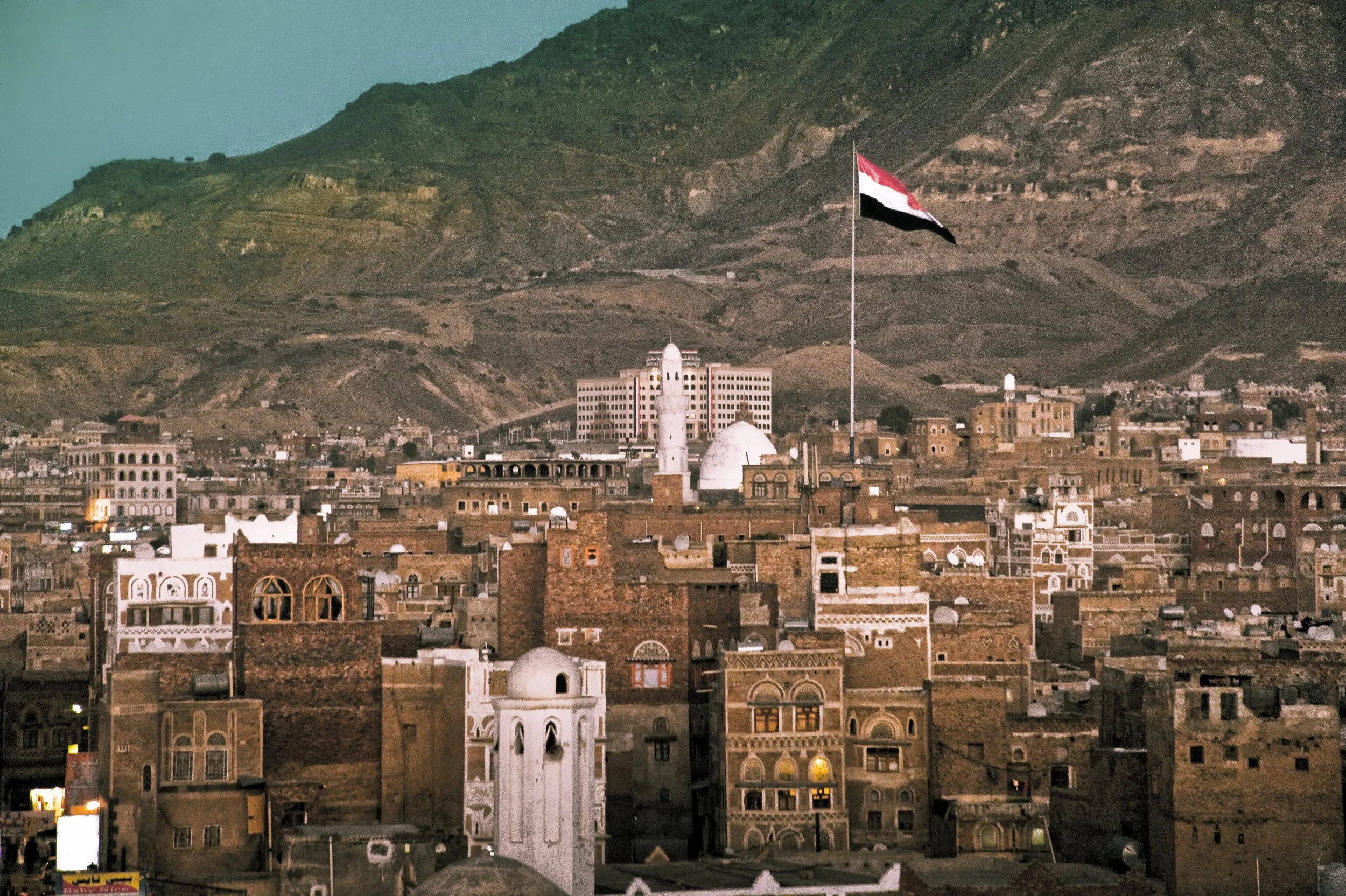 A cityscape with traditional Middle Eastern architecture, a large flagpole with a red, white, black, and green flag, and mountains in the background.