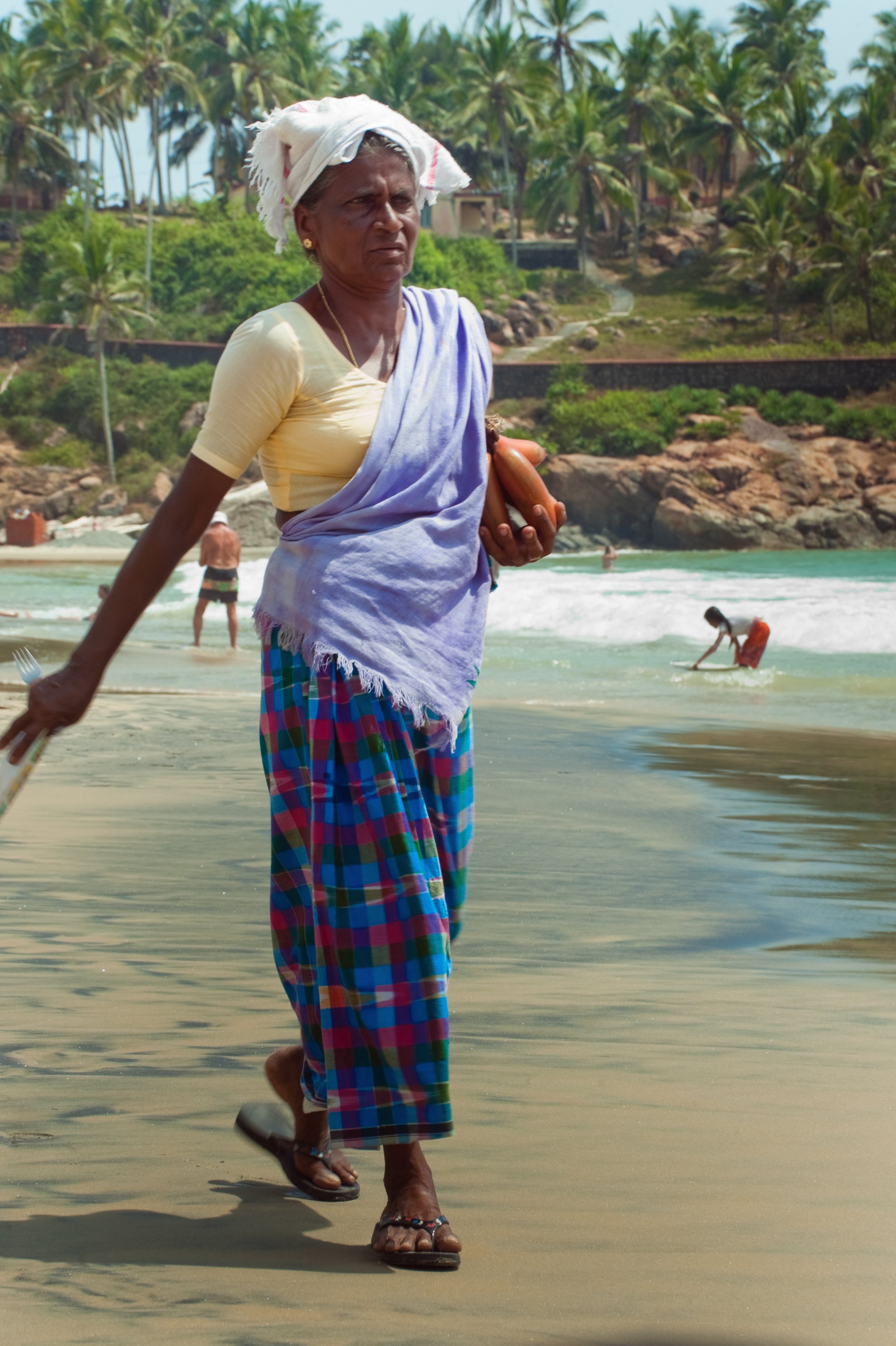 An elderly woman walking on the beach holding a vegetable, dressed in a yellow top, purple shawl, colorful checkered pants, and a headscarf, with palm trees and people in the background.