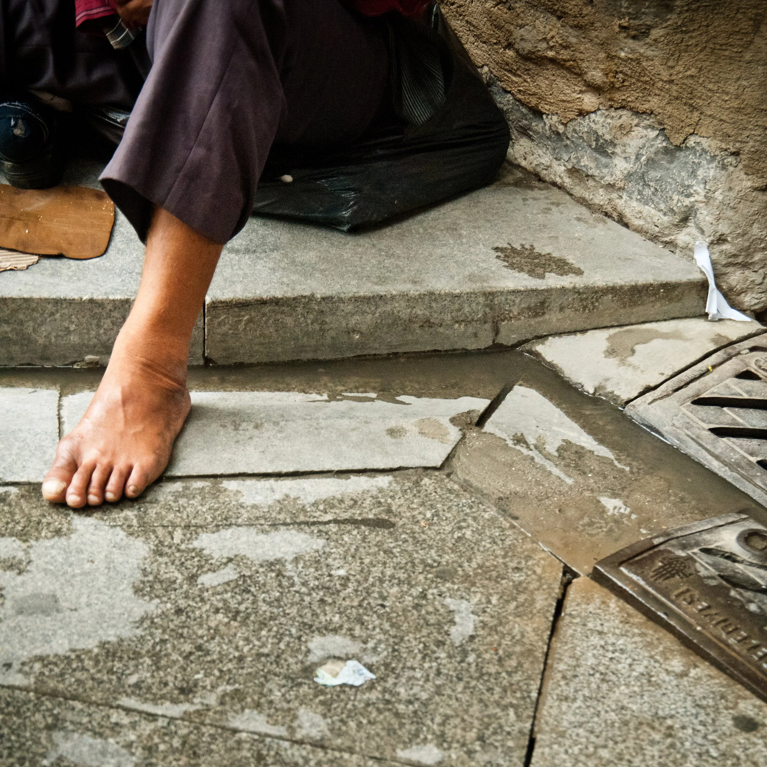 A person repairing or inspecting a sidewalk with broken and uneven tiles near a drainage grate.